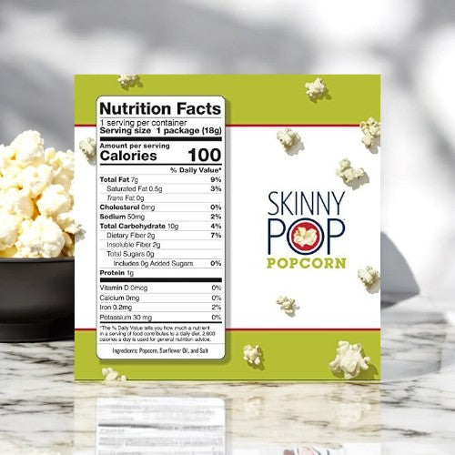 A pack of SkinnyPop Original Popcorn Snack Bags, 0.65 oz, displaying its nutritional information clearly on the front, sits invitingly. In the background, a small bowl filled with these guilt-free snacks made from whole-grain kernels tempts you to indulge.