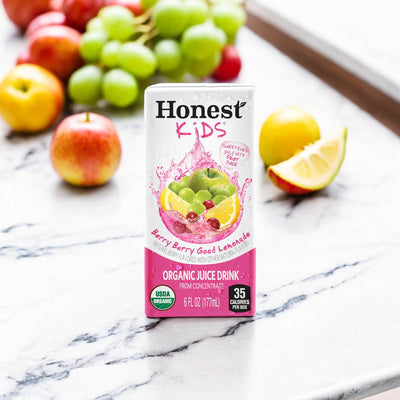 An Honest Kids Berry Good Lemonade - 6 oz - 1 Count carton is placed on a marble surface, surrounded by organic fruit juices including apples, grapes, and a sliced lemon. The carton emphasizes that it is USDA certified organic and contains only 35 calories per serving.