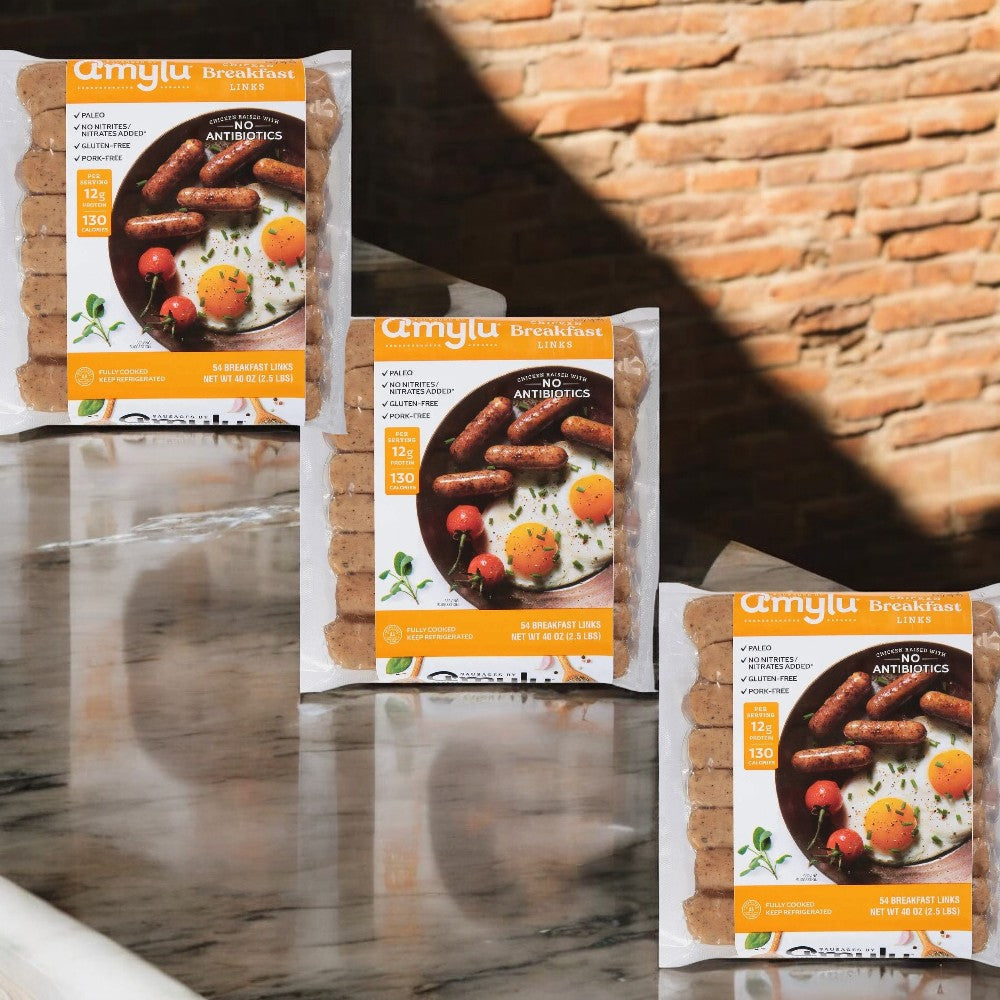 Three packages of Amylu Chicken Breakfast Sausage Links (40 oz) are showcased on a marble surface against a brick wall background, highlighting their all-natural ingredients.