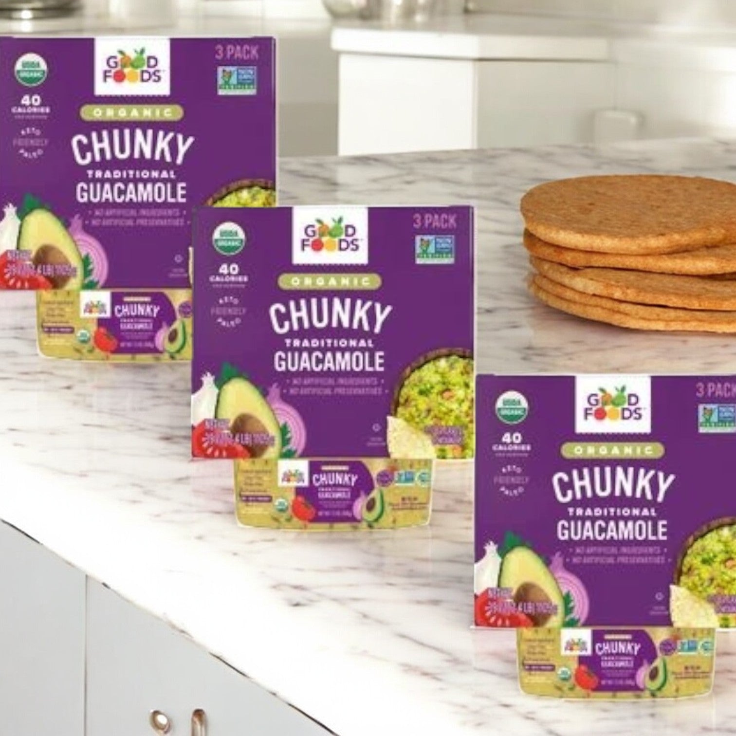 Three packs of Good Foods Organic Chunky Guacamole, praised for its chunky texture and non-GMO ingredients, are shown on a marble countertop with stacked cookies beside them.