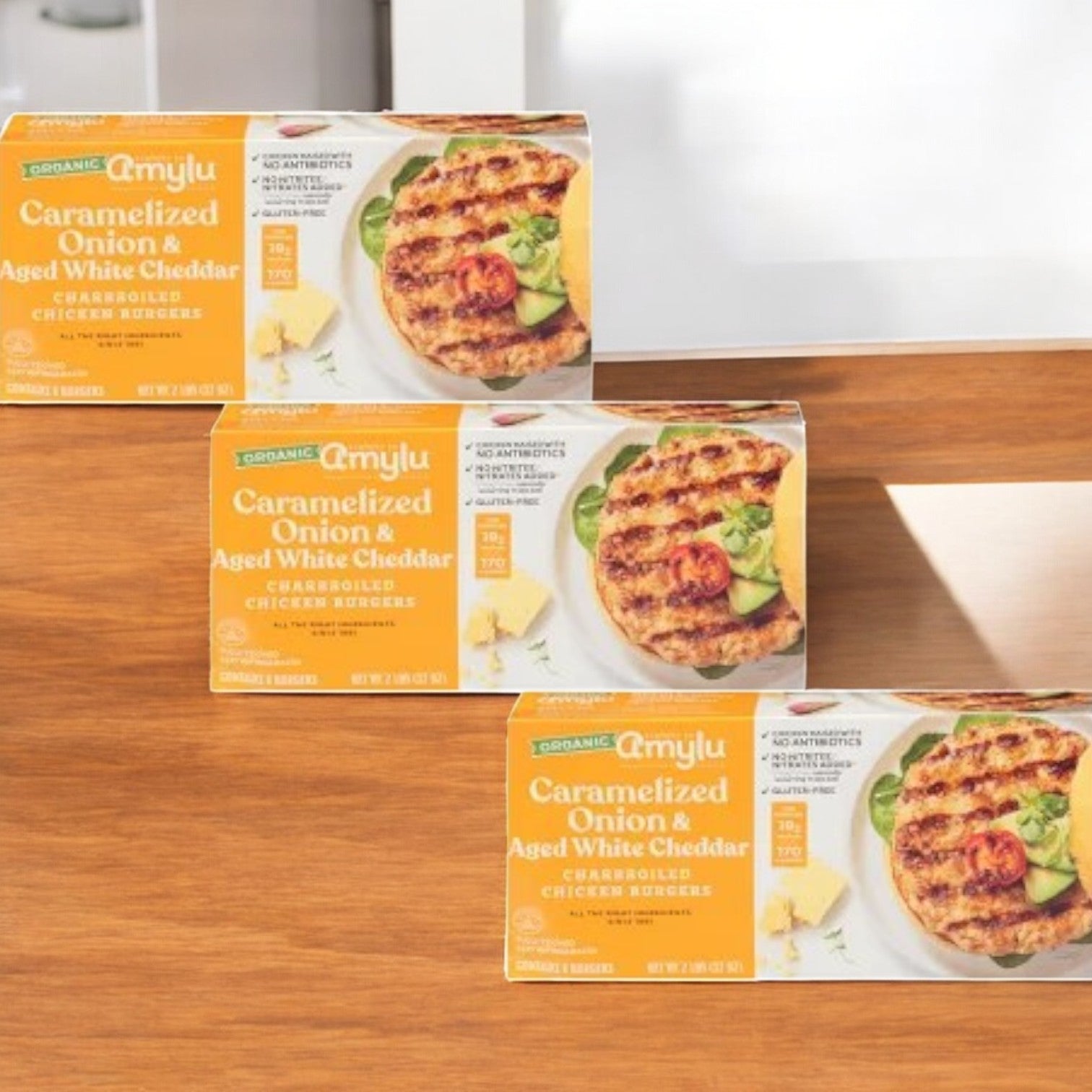 Three cases of Amylu Caramelized Onion and Aged White Cheddar Chicken Burgers, weighing 32 oz each and crafted from all-natural ingredients, are placed on a wooden surface.