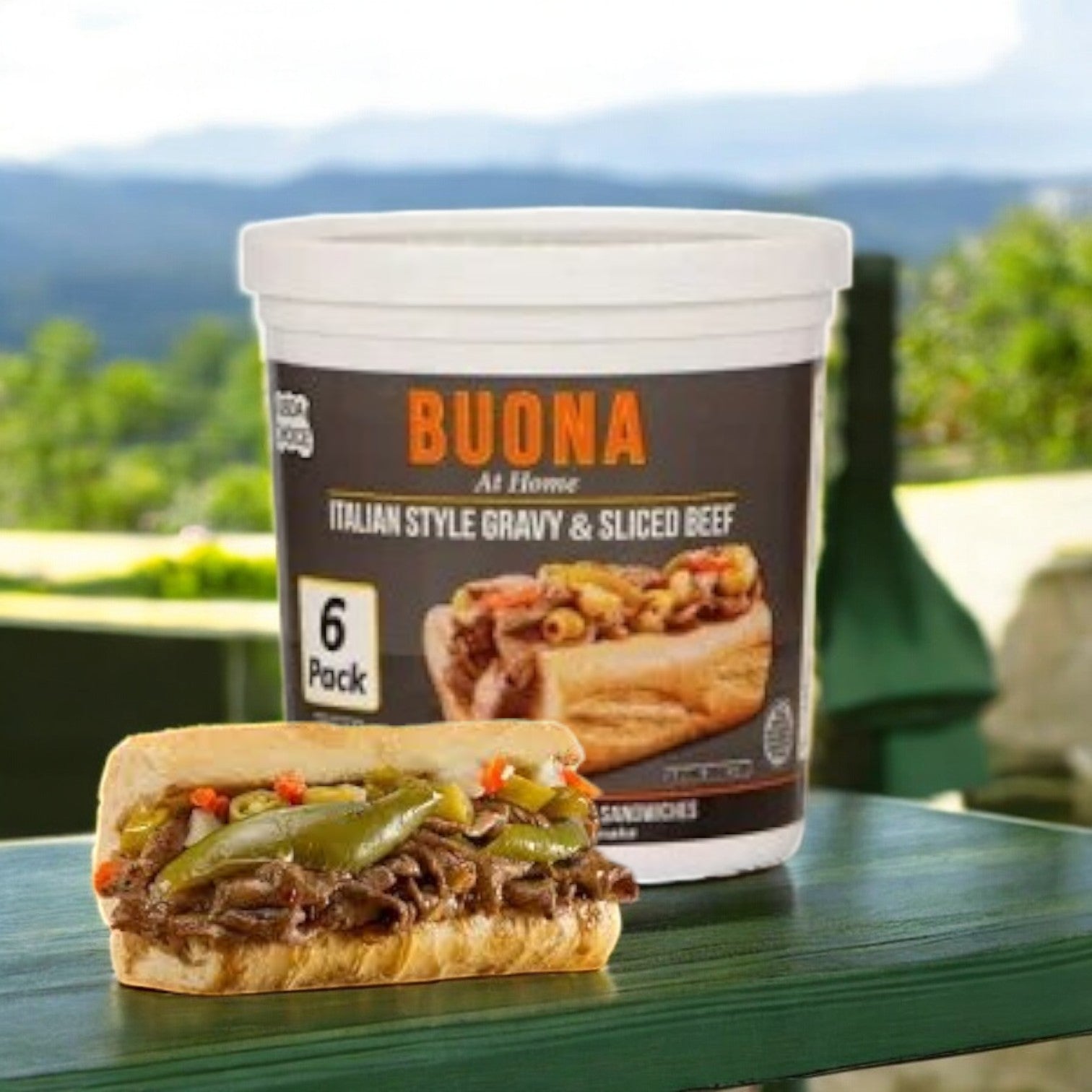 A Chicago-style treat awaits: Buona Italian Sliced Gravy & Italian Beef sits with a six-pack label behind an Italian beef sandwich topped with peppers on a green table. A scenic mountain view graces the background, enhancing the allure.