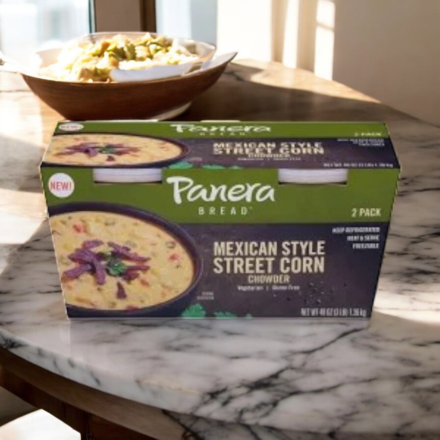 A package of Panera Bread Mexican Style Street Corn Chowder rests on a sunny marble table. In the backdrop, a bowl features roasted poblano peppers for a flavorful kick, reminiscent of classic elote.