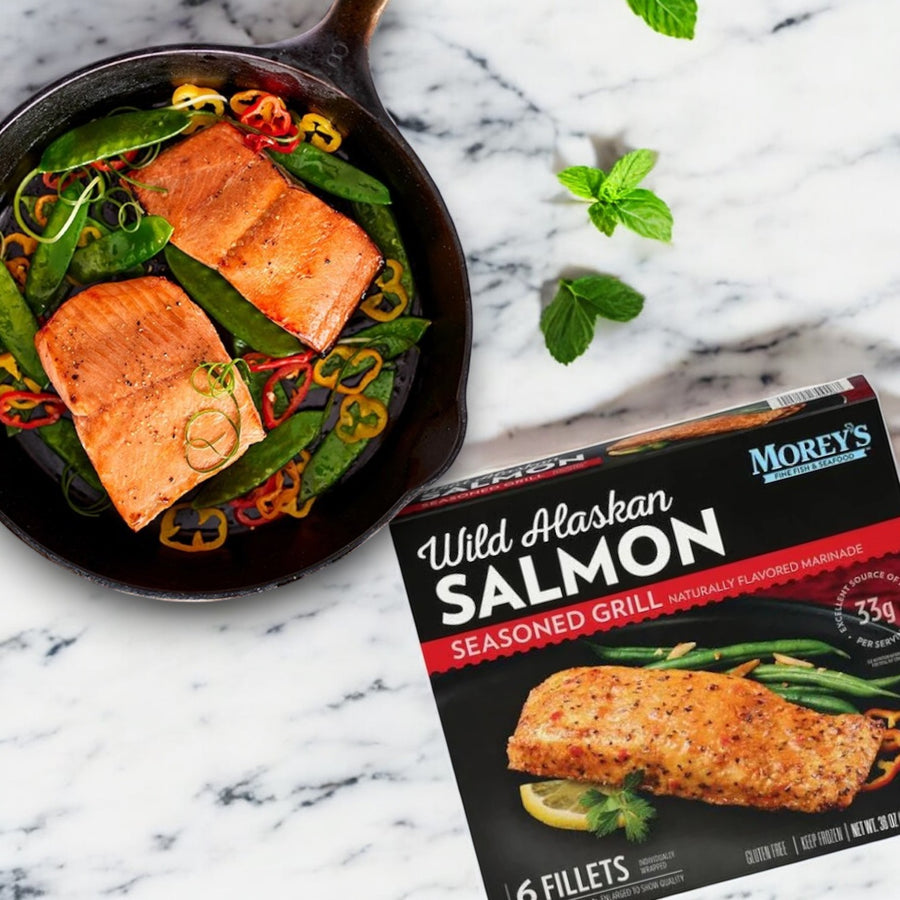 A skillet with cooked, marinated salmon, sliced peppers, and green beans sits enticingly next to a box of Morey's Marinated Wild Alaskan Salmon, 6 ct by Morey's Fine Fish & Seafood on a marble surface.
