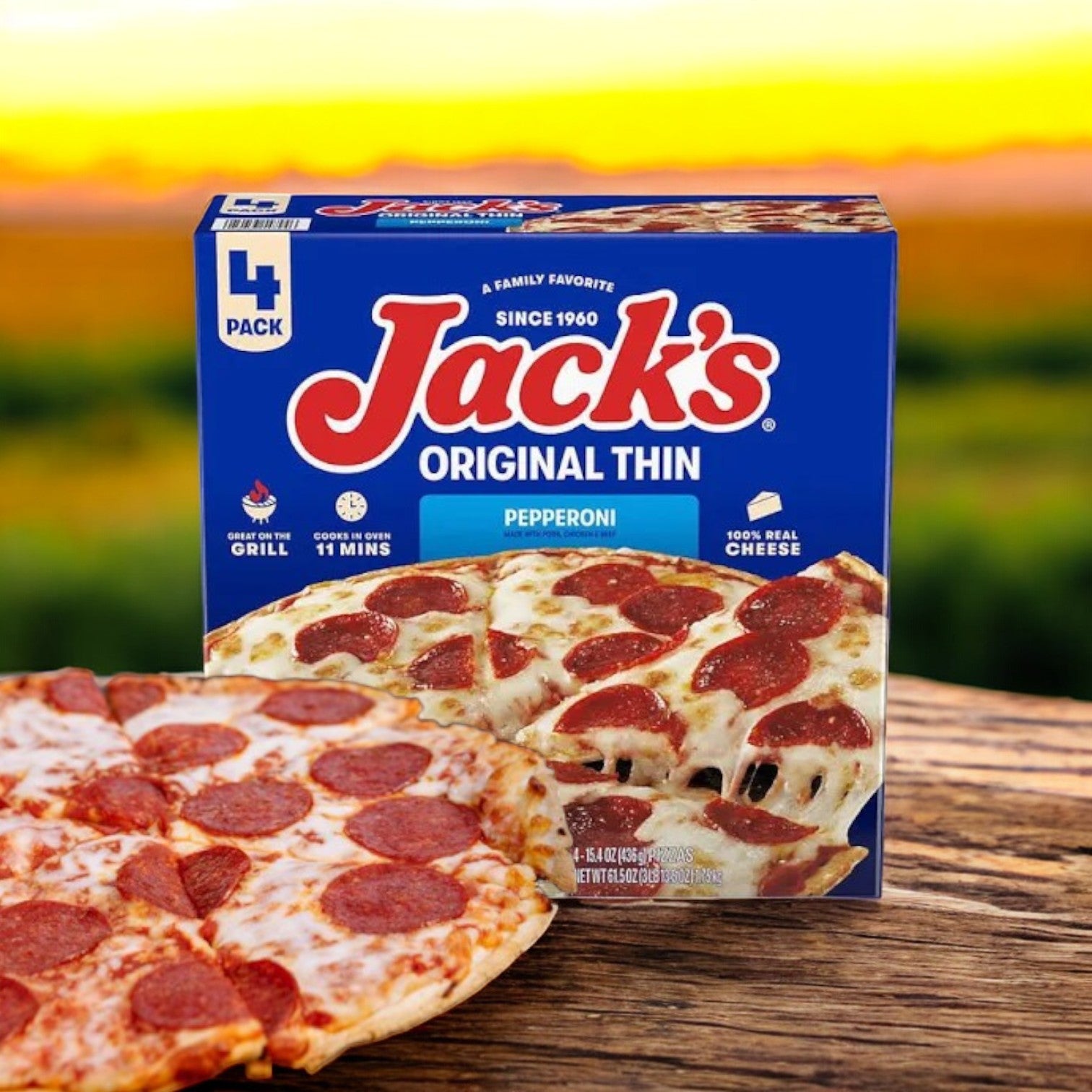 A box of Jack's Original Thin Pepperoni Frozen Pizza, from a 3-pack set, sits beside a gooey mozzarella-covered pizza on a wooden table. The blurred outdoor backdrop emphasizes the delicious simplicity of this delightful frozen pizza offering from Jack's.