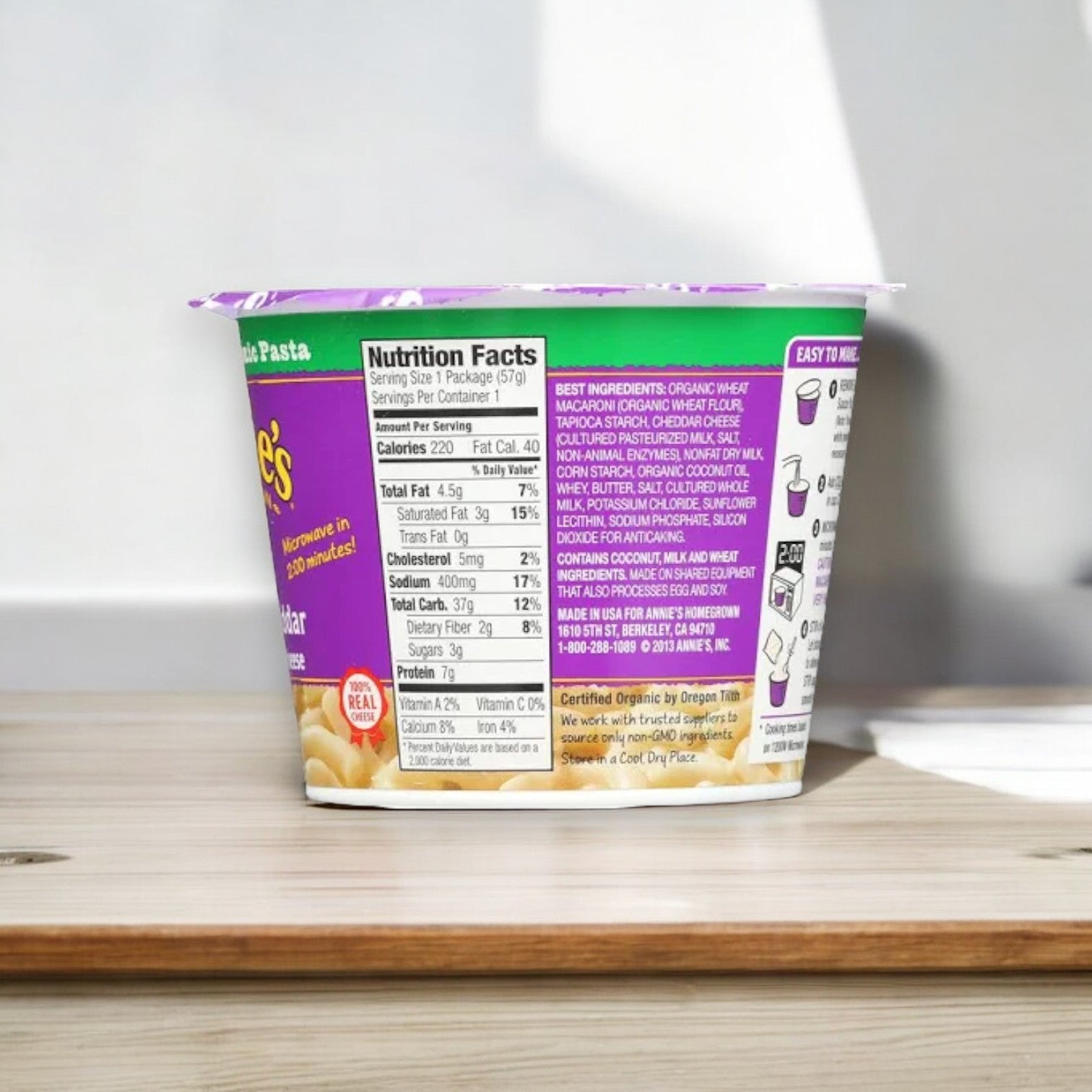 A cup of Annie's White Cheddar Mac and Cheese Cups, 2.1 oz - 1 Count by Annie's, placed on a wooden surface, showcasing the nutrition facts and ingredients on the side.