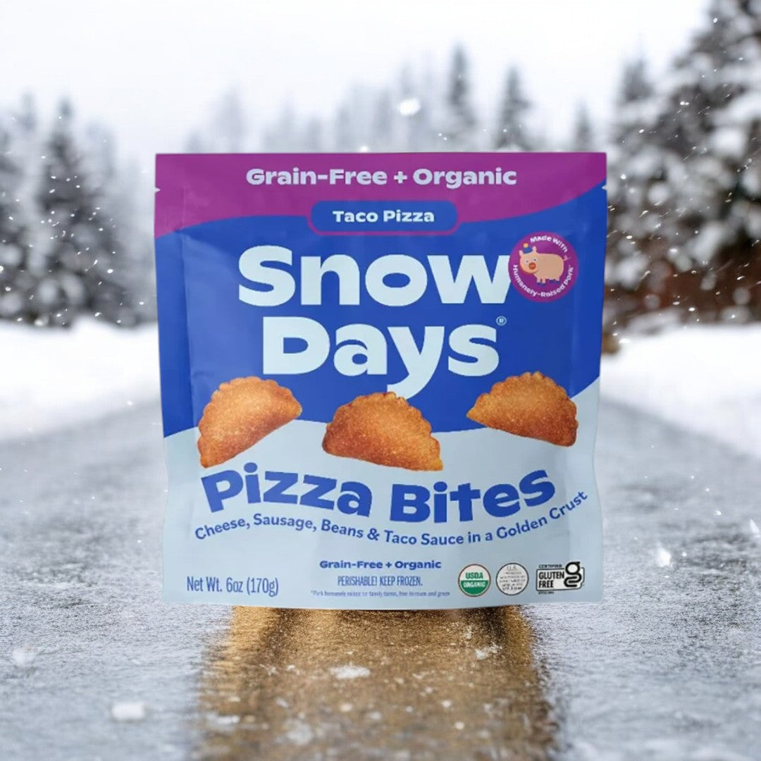 A bag of Snow Days Frozen Organic Taco Pizza Bites, 6oz.- 1 Count, displayed against a snowy road scenery. The packaging emphasizes "Grain-Free + Organic" and features the distinctive blend of cheese, sausage, beans, and taco sauce encased in a golden crust.