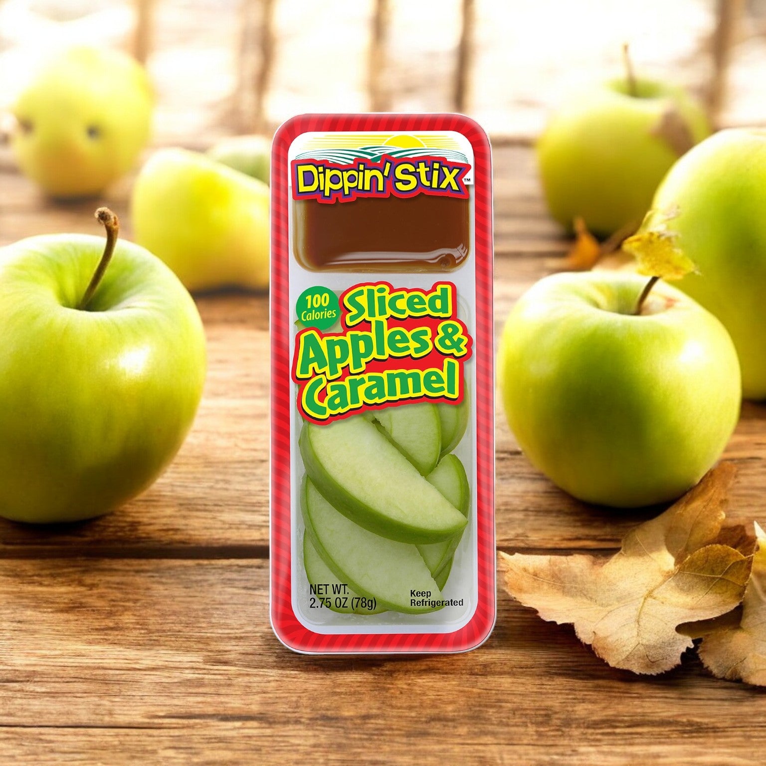 A package of Dippin' Stix Tart Apples & Caramel Snack Packs (2.75 oz, 1 count) is placed in front of whole green apples and a few dry leaves on a wooden surface.