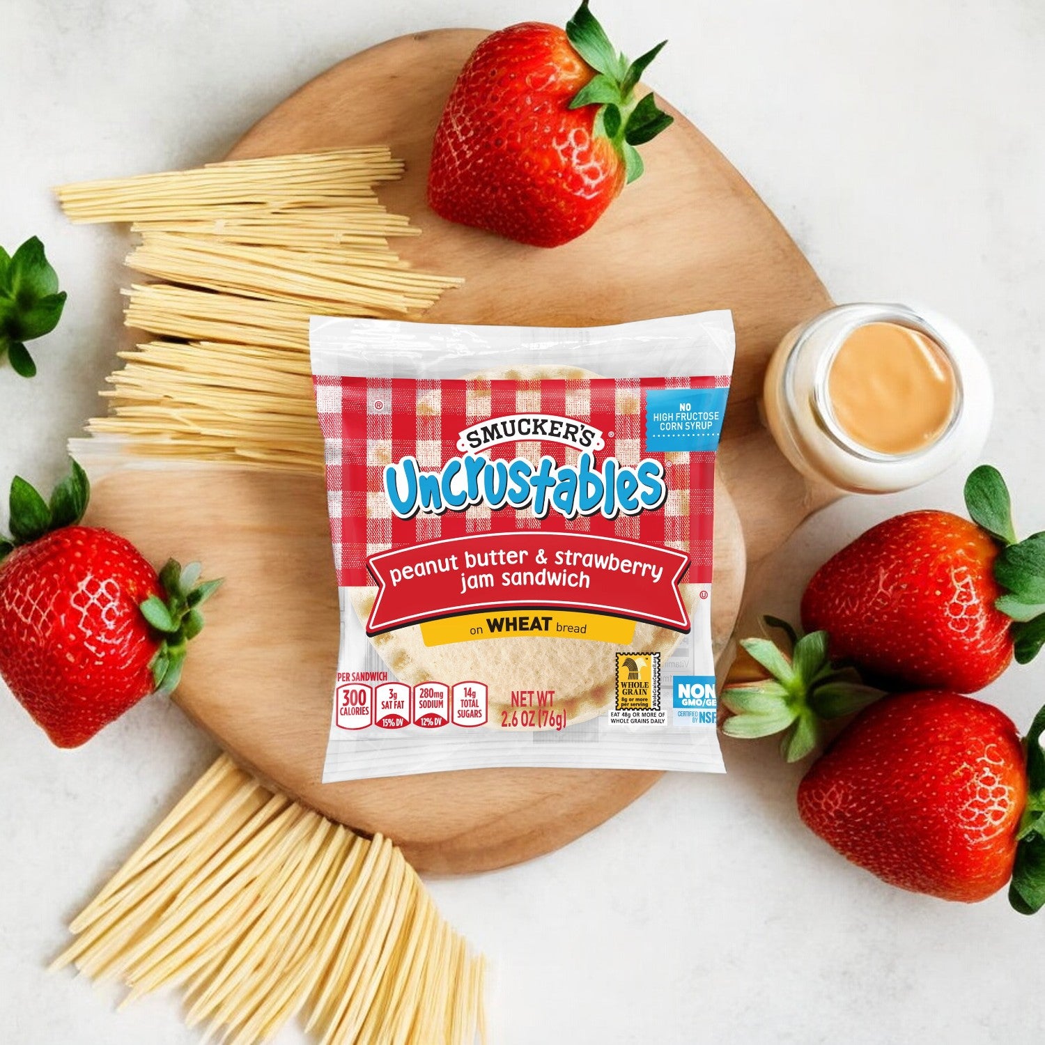 An Uncrustable Smuckers Peanut Butter & Strawberry Jelly sandwich packet sits on a wooden cutting board alongside fresh strawberries, uncooked spaghetti, and a small jar of creamy peanut butter on a white background.