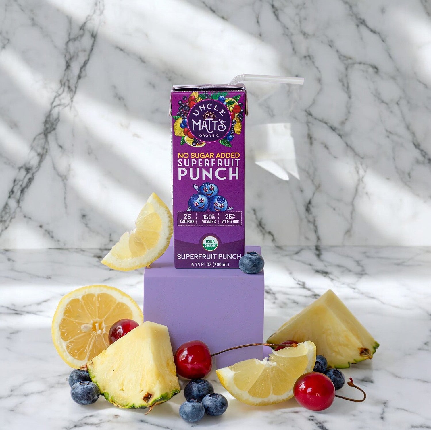 A carton of Uncle Matt's Superfruit Punch Juice Box, 6.75 oz, sits on a marble surface surrounded by blueberries, pineapple slices, cherries, and lemon slices—perfect for those seeking convenient portable drinks.