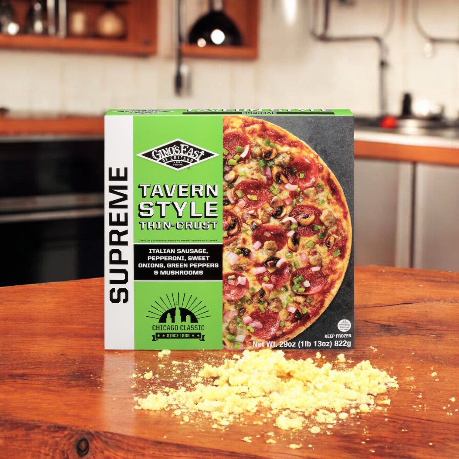 A box of Gino's East Supreme Thin-Crust Tavern Style Pizza, weighing 29 oz., is displayed on a wooden surface in a kitchen. Some crumbled cheese is spilled in front of the box, adding to the classic look.