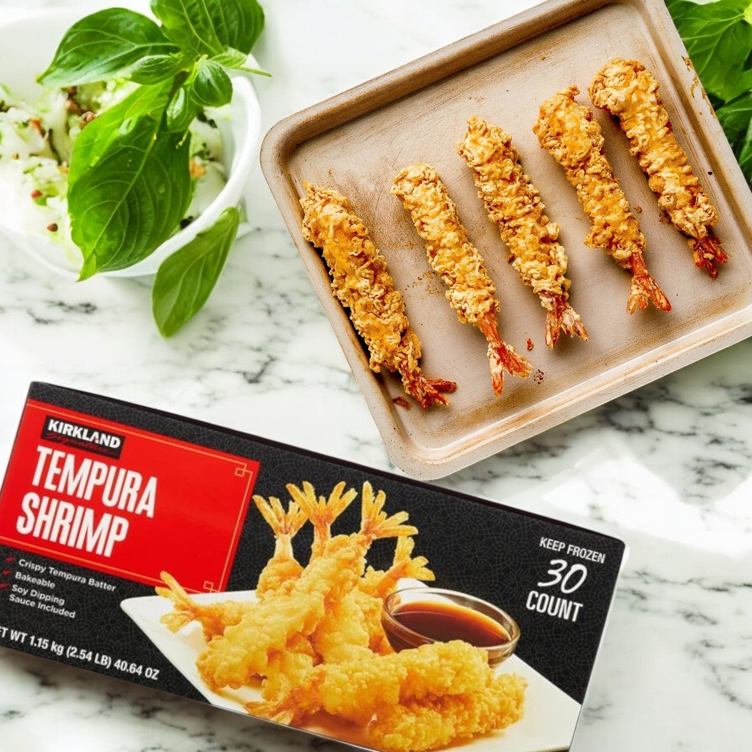 A baking tray with five crispy tempura shrimp from Easy Lunches Kirkland Signature Tempura Shrimp, 2.35 lbs, 30-count - 3 Packs sits beside its box on a marble counter, with a bowl of greens in the background.