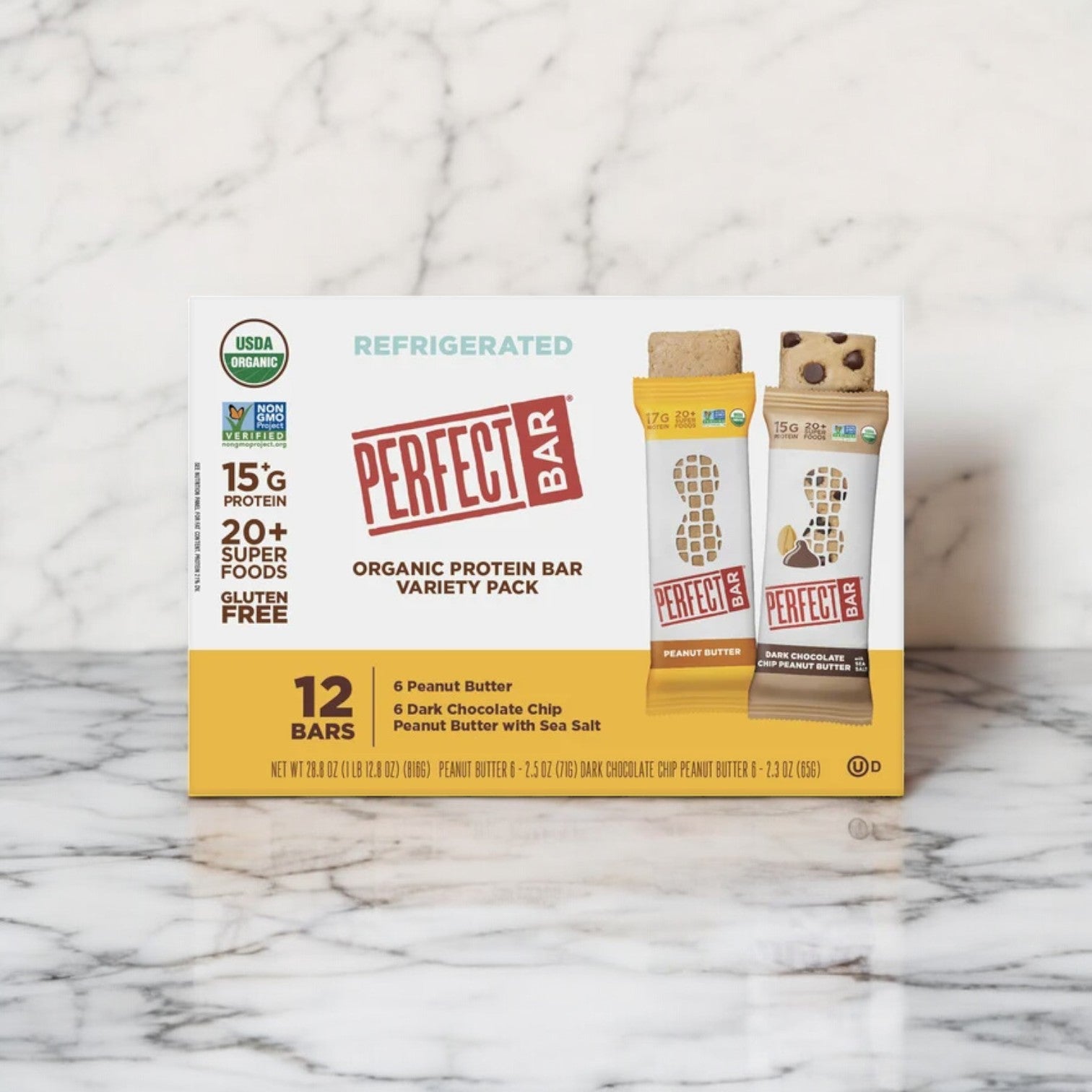 A box of Perfect Bar Refrigerated Organic Protein Bars is displayed gracefully on a marble surface. This variety pack contains 24 bars, featuring the delightful flavors of dark chocolate chip peanut butter and peanut butter. Indulge in a nutritious treat packed with superfoods in every bite from Perfect Bar.