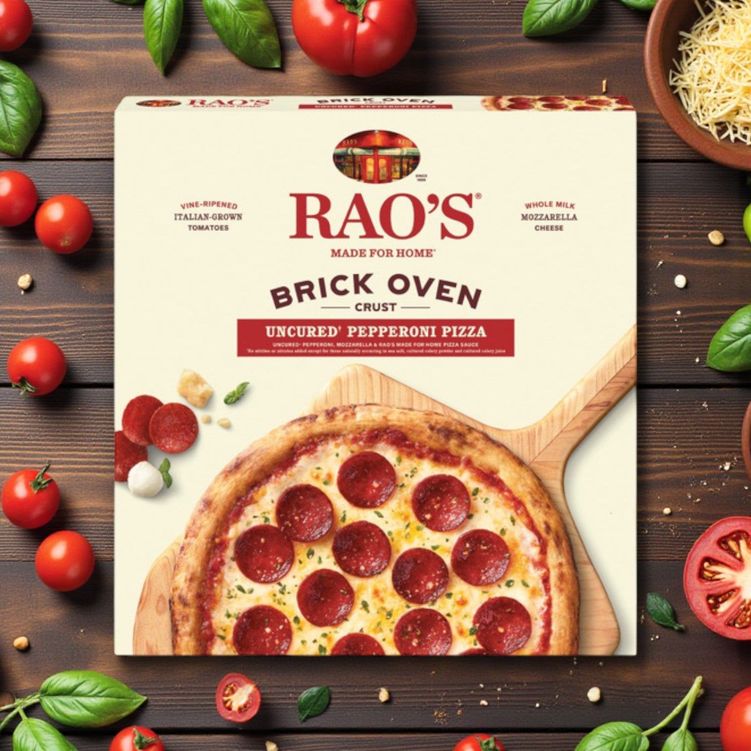 A box of Rao's Uncured Pepperoni Brick Oven Crust Pizza – 18.3 oz sits on a wooden table, surrounded by tomatoes, basil, grated cheese, and a sliced pizza topped with uncured pepperoni.