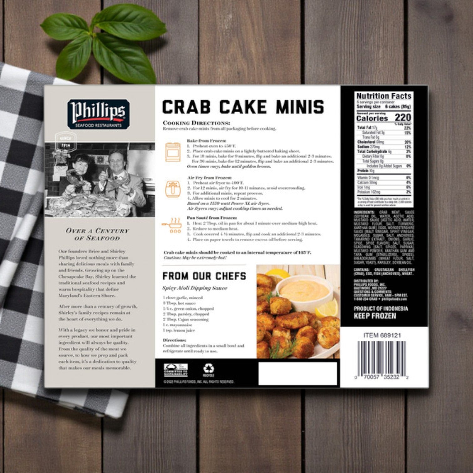 A box of Phillips Crab Cake Minis – 36 Count (1 lb 2 oz) by Phillips Foods sits on a wooden surface beside a checkered napkin and basil; packaging shows cooking directions—perfect seafood appetizers for any occasion.