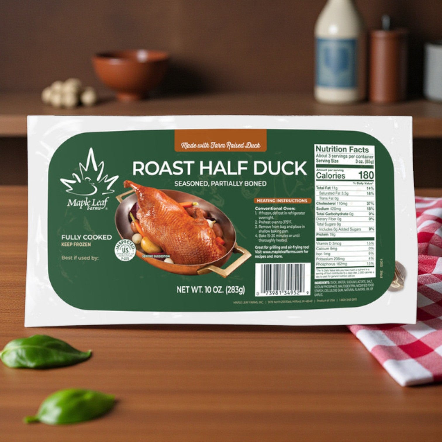 The Maple Leaf Farms Half Roast Duck – 10 oz, fully cooked and seasoned, is shown on a wooden table with basil and kitchen décor—an easy gourmet entrée at home from Maple Leaf Farms.