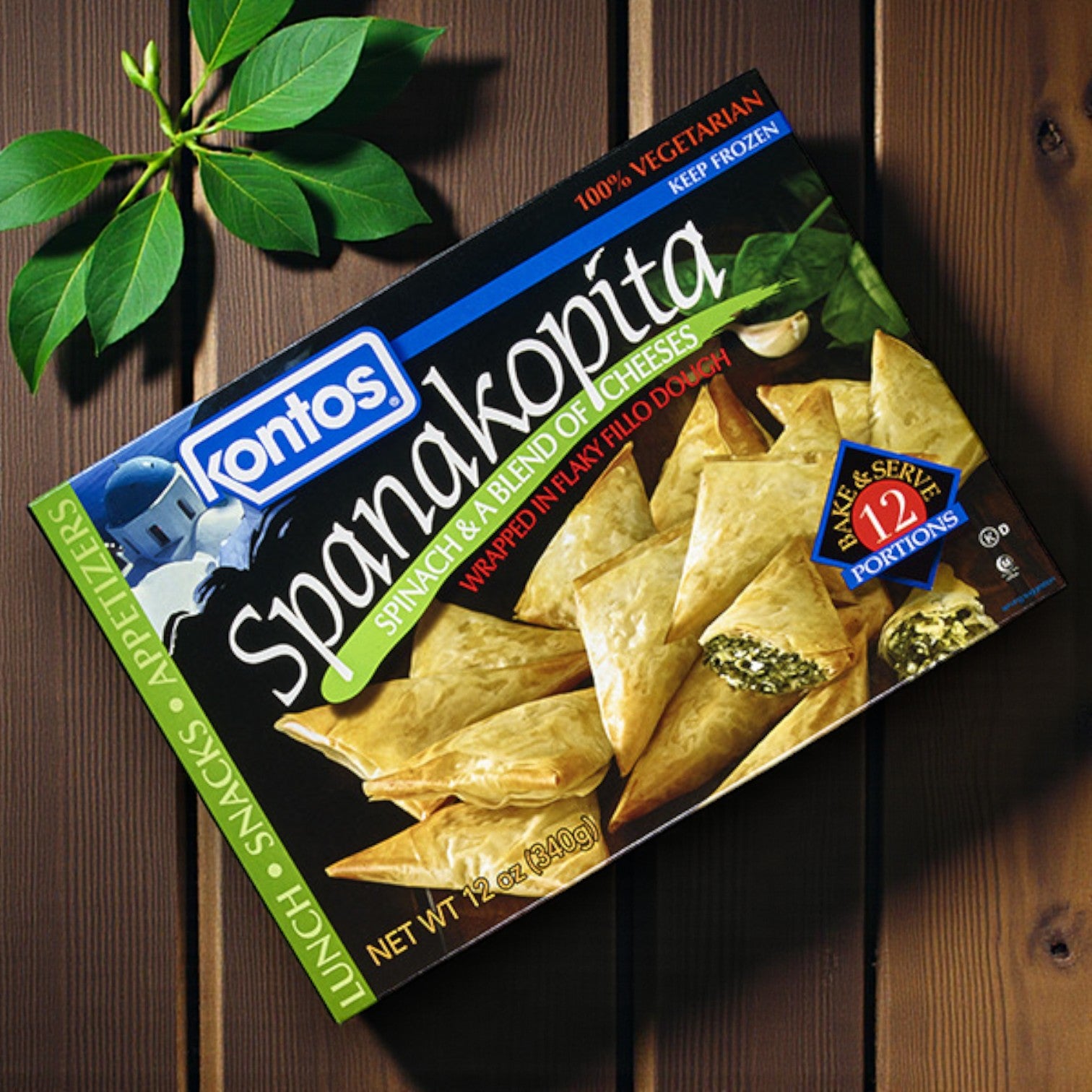 A box of Easy Lunches Kontos Spanakopita – 12 Count, a vegetarian Greek appetizer with spinach and feta, sits on a wooden surface next to green leaves.