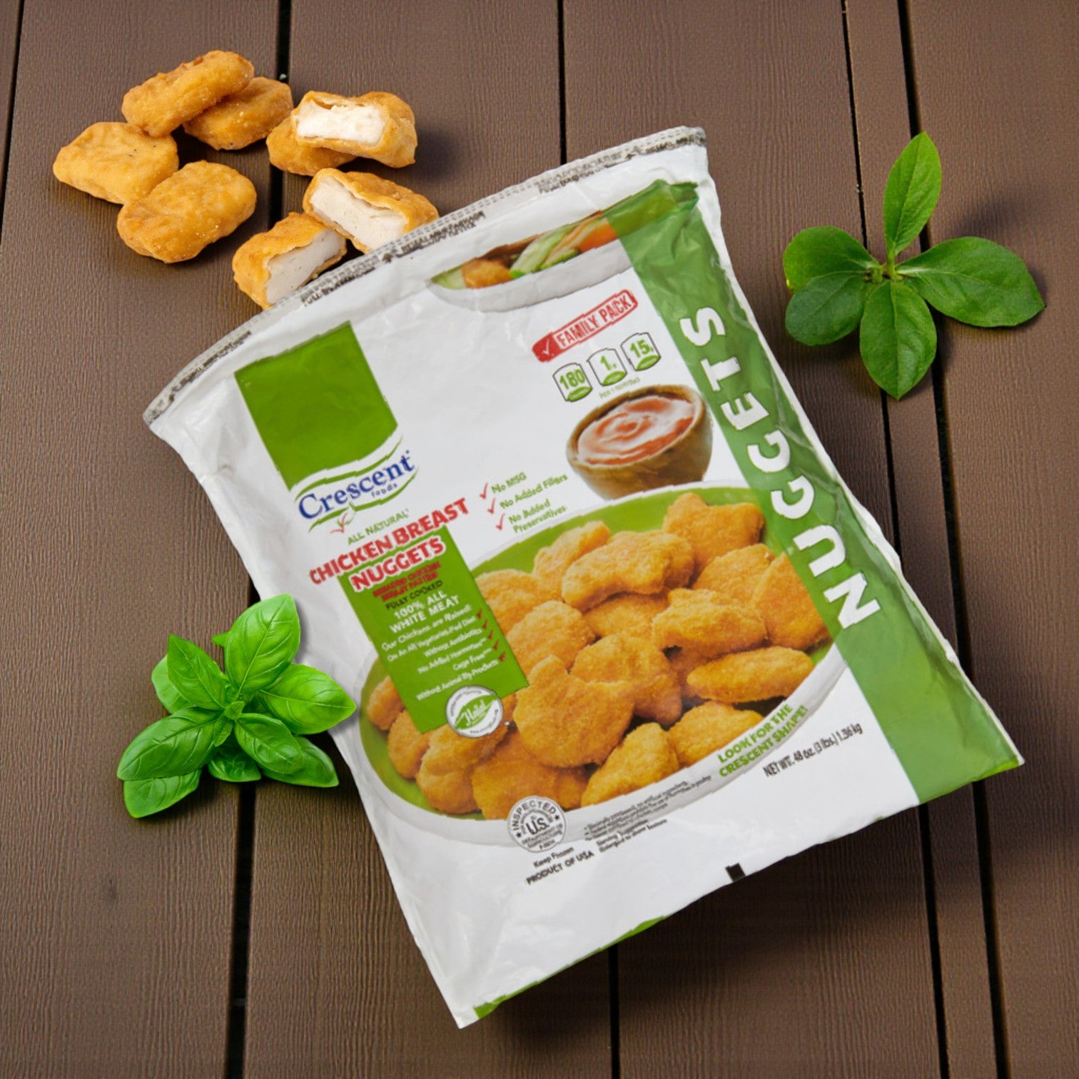 A 3 lb bag of Crescent Foods Halal Chicken Breast Nuggets sits on a wooden surface, surrounded by several nuggets, fresh basil leaves, and a small cup of dipping sauce.