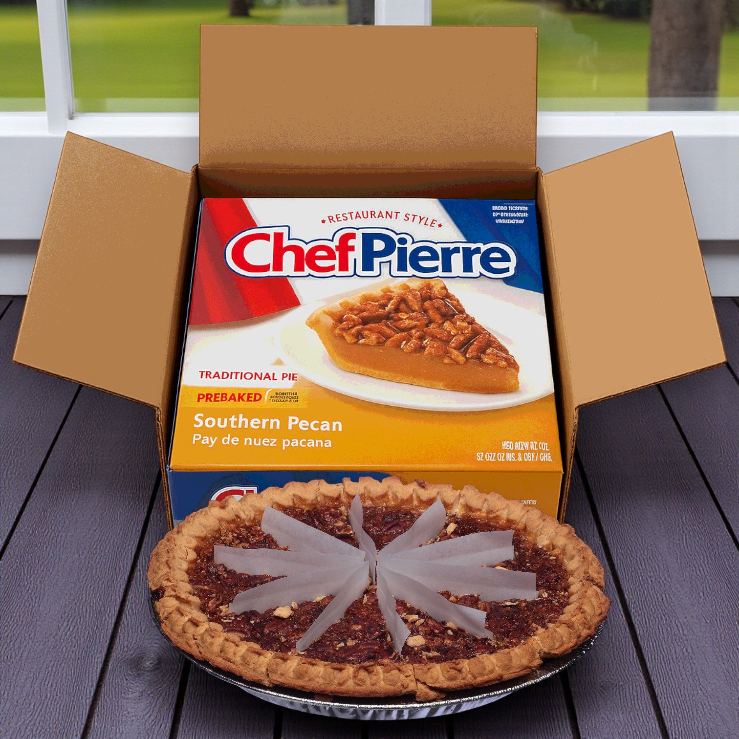 An open box of Chef Pierre Pre-Baked Southern Pecan Pie, 36 oz. - 3 Count sits on a table, with one pre-baked pecan pie in front of it topped with several pieces of clear plastic.