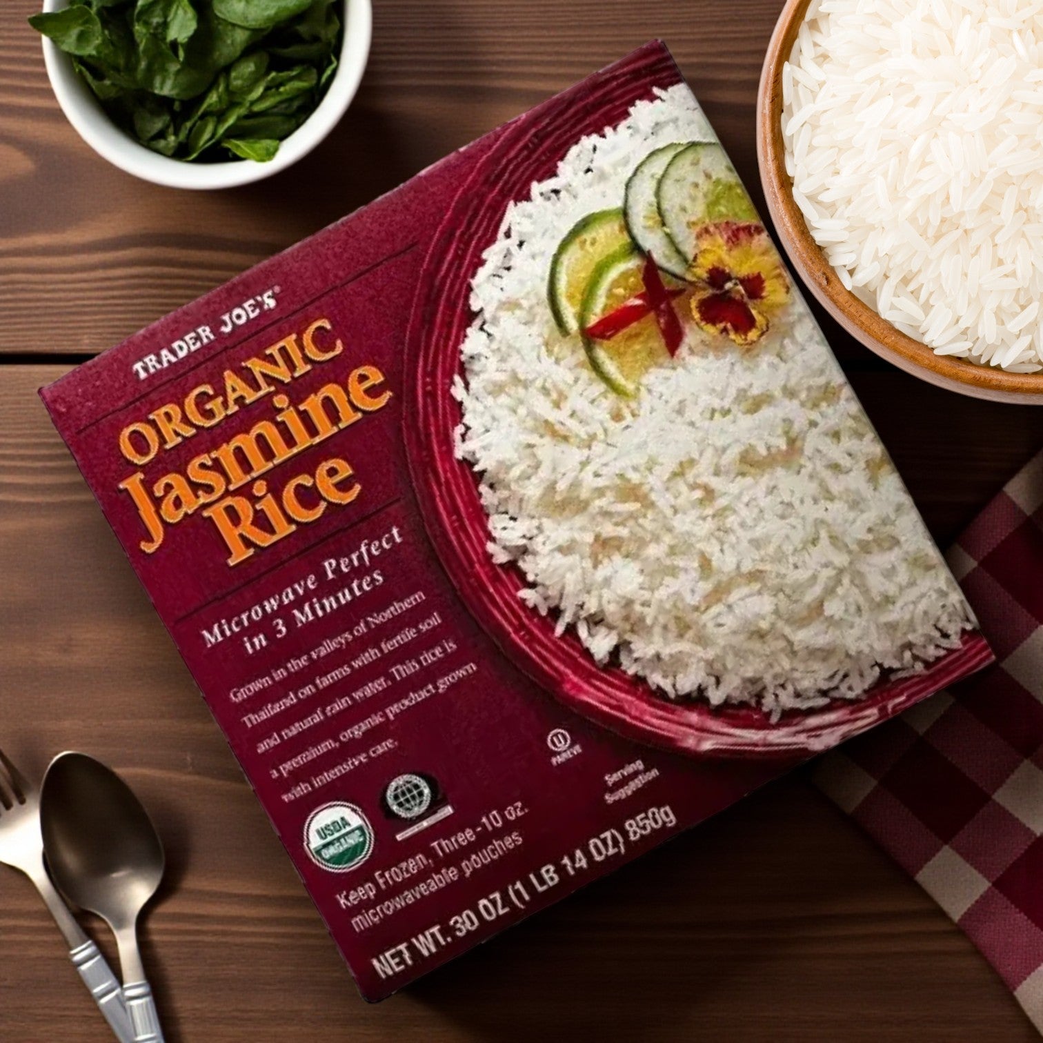 A box of Easy Lunches Trader Joe’s Organic Jasmine Rice, 30 oz – 3 Pack is on a table beside a bowl of Thai Jasmine Rice, fresh spinach leaves, utensils, and a red checkered cloth.