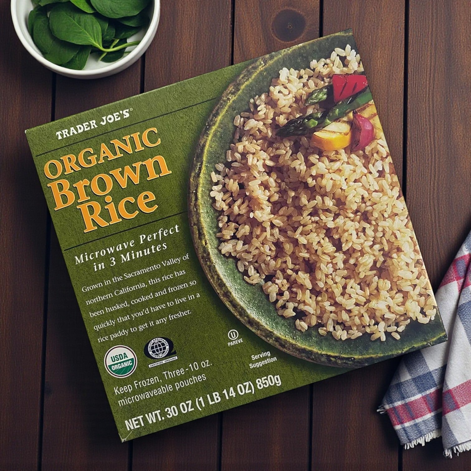 A pack of Easy Lunches Trader Joe’s Organic Brown Rice, 30 oz – 3 Pack, sits on a wooden table beside a bowl of spinach and a cloth napkin—microwave-ready rice for quick, wholesome meals.