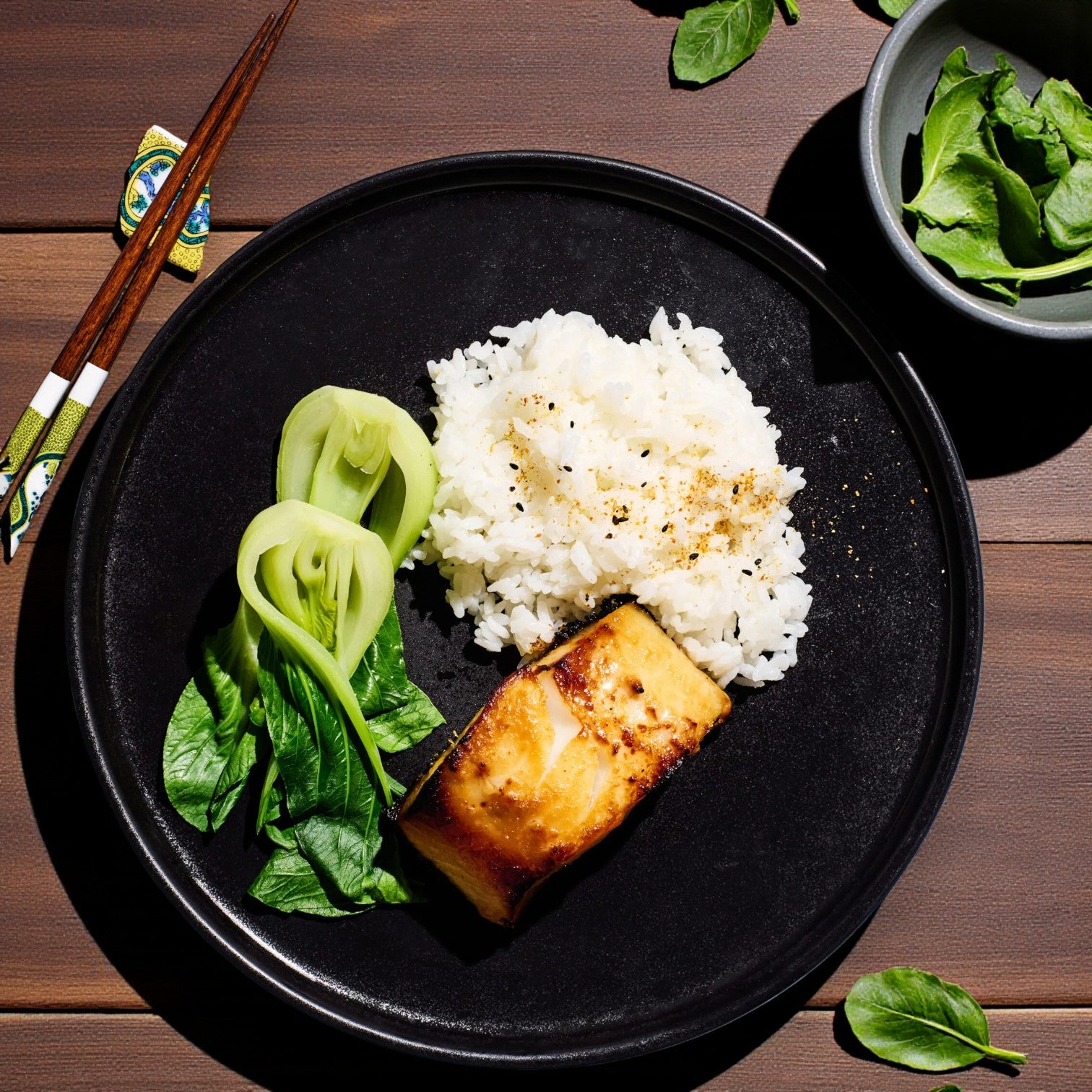 A black plate with white rice, a glazed Alaskan Leader Seafoods Alaska Cod Fillet, Wild Caught, 2lbs., and steamed bok choy sits beside chopsticks and a bowl of fresh greens on a wooden table.