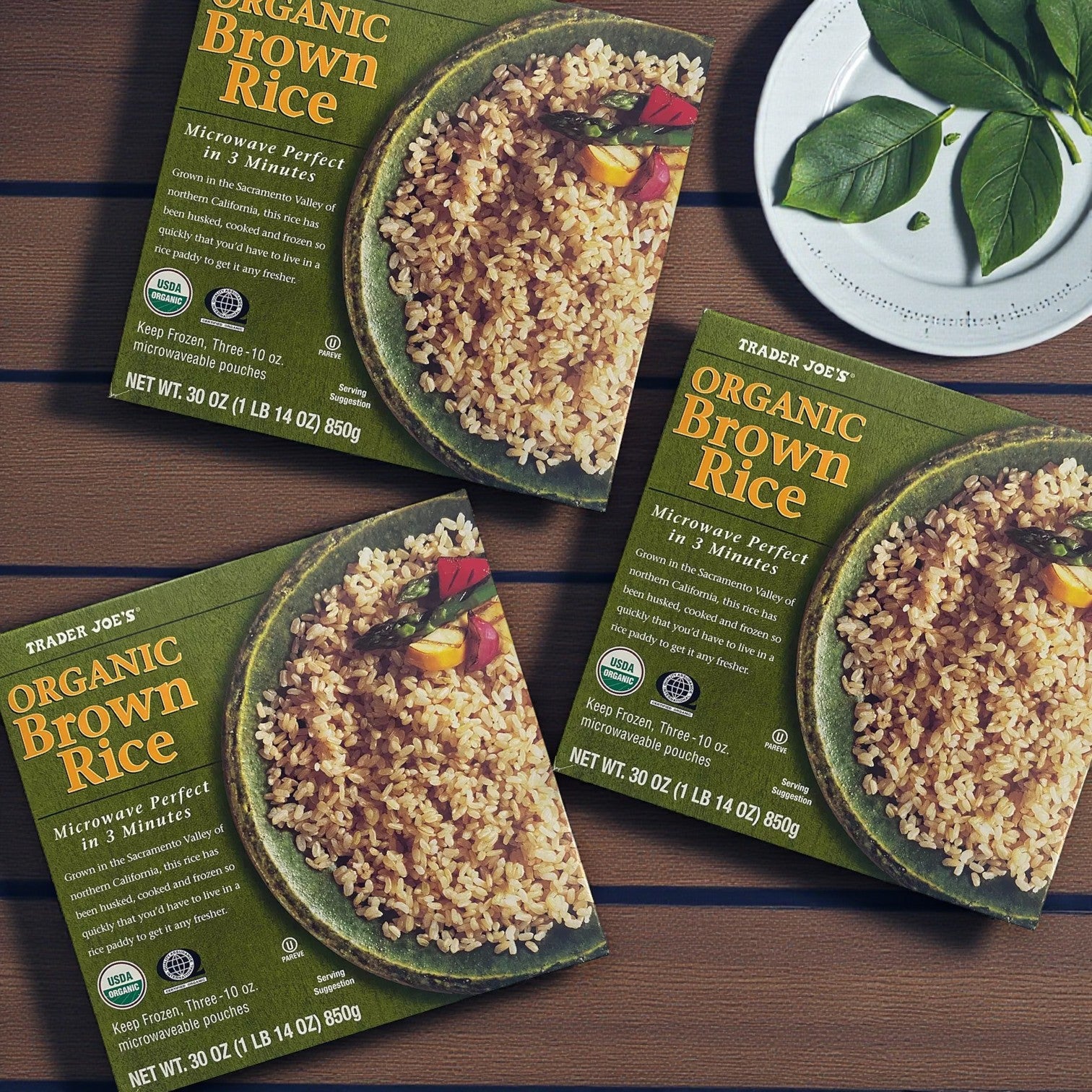Three Easy Lunches Trader Joe’s Organic Brown Rice, 30 oz – 3 Pack boxes are arranged on a wooden surface beside a white dish with green leaves, showcasing this easy microwave-ready rice option.