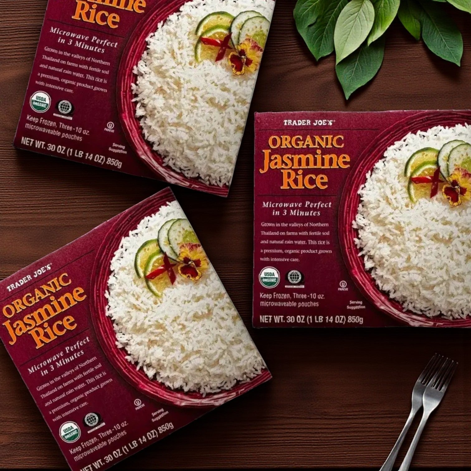 Three packs of Easy Lunches Trader Joe’s Organic Jasmine Rice (30 oz) are displayed on wood beside green leaves. The microwave-ready packaging features rice garnished with cucumber, chili, and herbs.