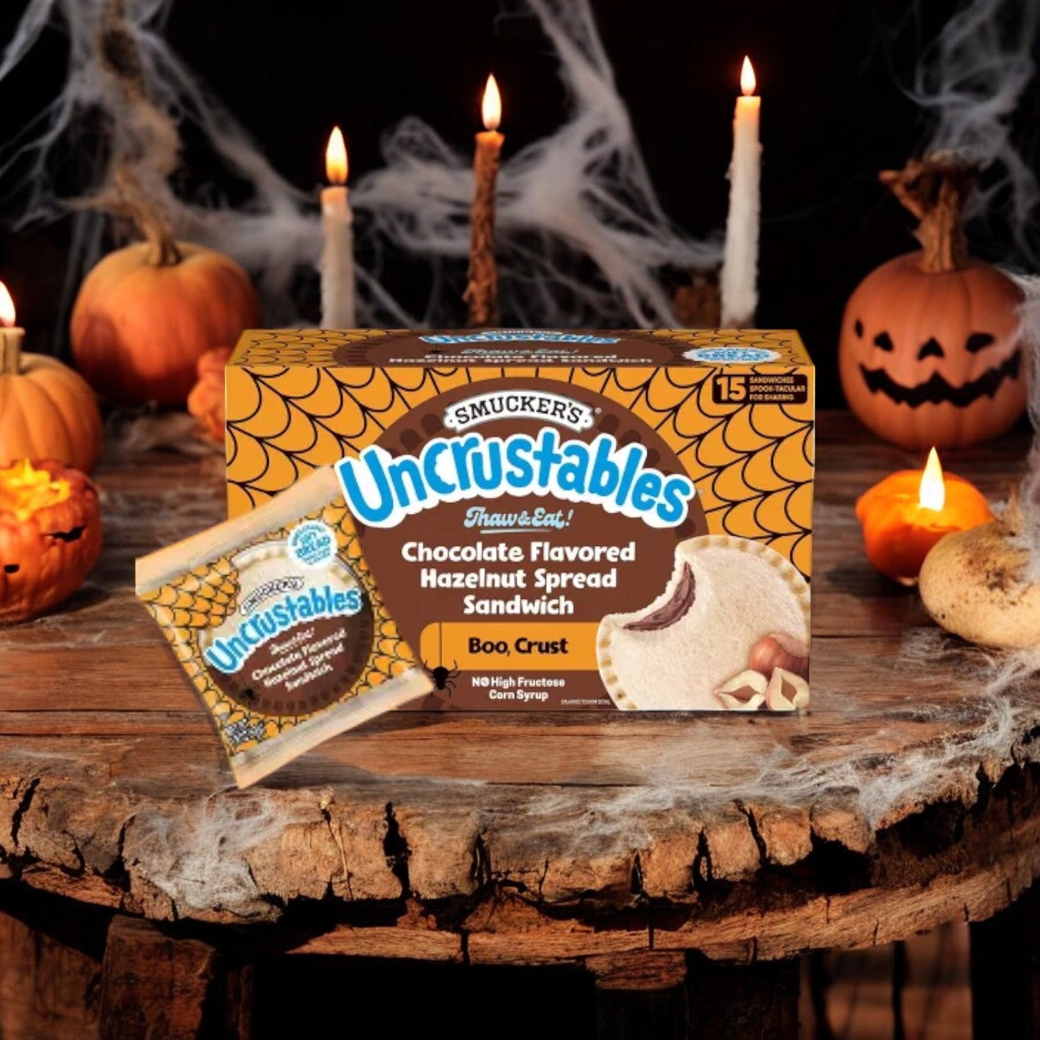 A box of Uncrustable Smuckers Sandwiches, Chocolate Flavored Hazelnut Spread Sandwich, 15 Count, 2 Pack named "Boo Crust," perfect for on-the-go snacking, placed on a wooden table with pumpkins, candles, and spider webs in the background.