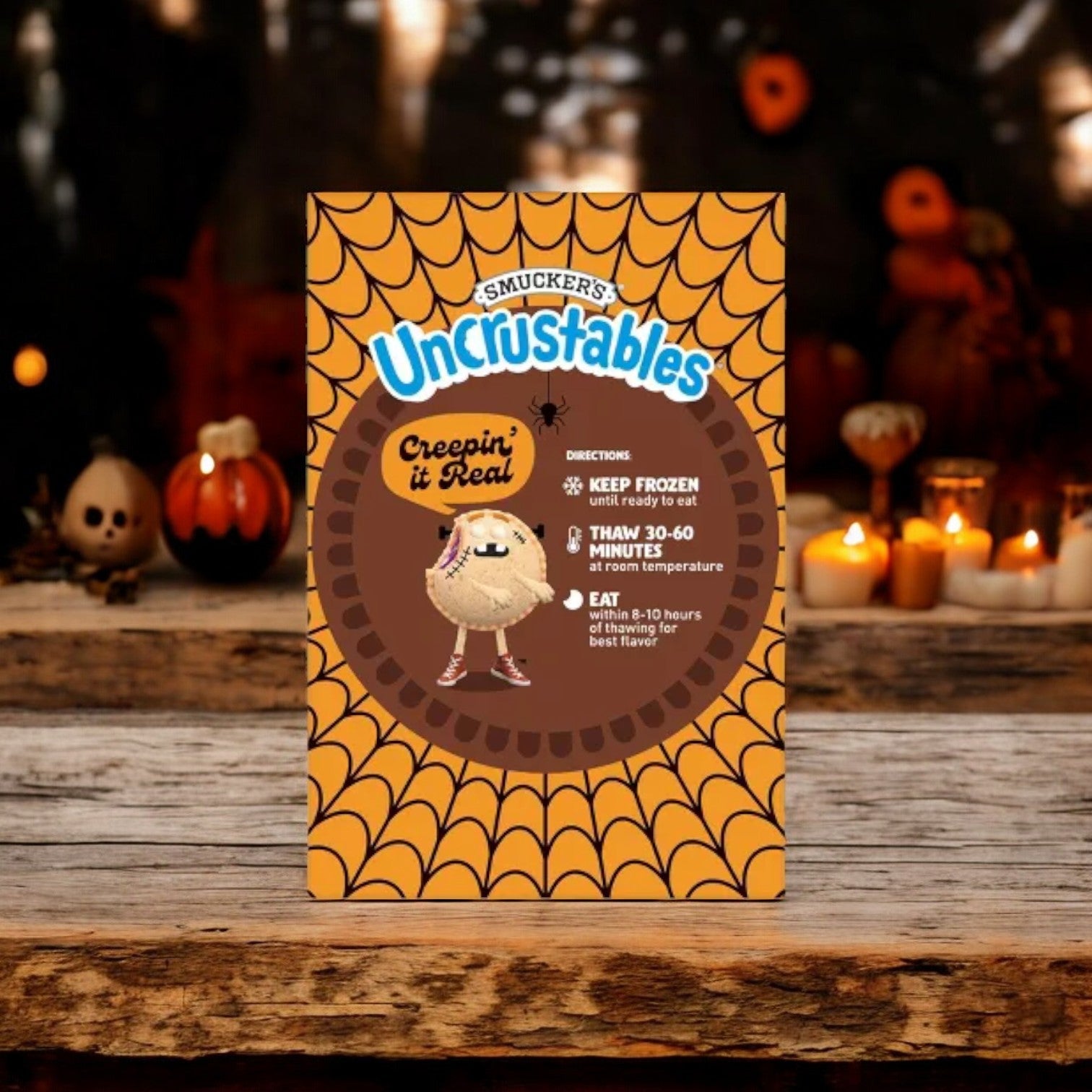 A box of Uncrustable Smuckers Sandwiches with a Halloween-themed design sits on a wooden table adorned with pumpkins and candles. These Smucker's Sandwiches, featuring chocolate-flavored hazelnut spread, come individually wrapped in a convenient 15 count, 2 pack—perfect for on-the-go snacking. For best results, keep them frozen and thaw for 30-60 minutes or overnight in the fridge before enjoying.