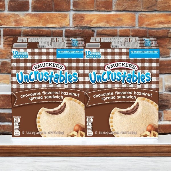 Two boxes of Uncrustable Smucker's Sandwich Chocolate Flavored Hazelnut Spread Sandwiches are displayed against a brick wall background. Each box contains ten individually wrapped 1.8 oz sandwiches, making a total of 20 sandwiches perfect for an on-the-go snack.