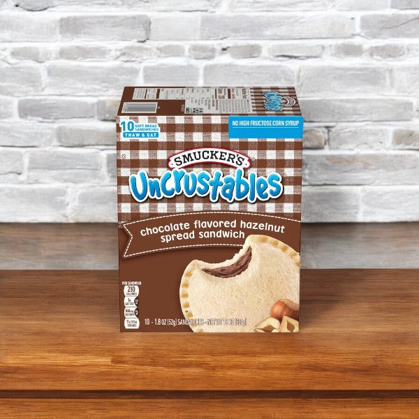 A 2-pack box of Uncrustable Smuckers Sandwiches, featuring chocolate-flavored hazelnut spread and totaling 20 sandwiches, sits on a wooden surface in front of a white brick wall, perfect for an on-the-go snack.