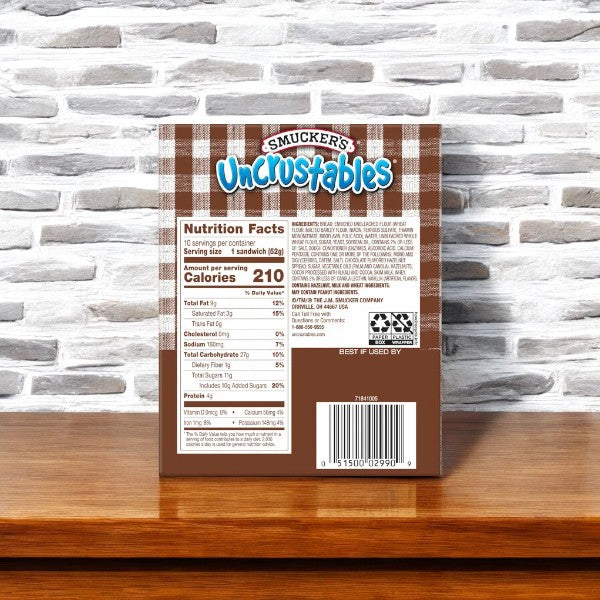 The image shows the back of an Uncrustable Smucker's Sandwich box, specifically the Chocolate Flavored Hazelnut Spread Sandwiches, displaying nutrition facts and ingredients against a brick wall background. Highlighting their practicality, these individually wrapped sandwiches make for a perfect on-the-go snack. The box contains 10 sandwiches per pack, with a total of 20 sandwiches in this 2-pack offering.