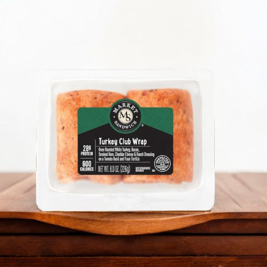 A packaged E.A. Sween Turkey Club With Bacon, Ham, Cheddar Cheese, & Ranch Dressing Wrap is placed on a wooden surface. The label highlights 29g protein, 600 calories, and includes details like smoked bacon, cheddar cheese, ranch dressing, and a flour tortilla.