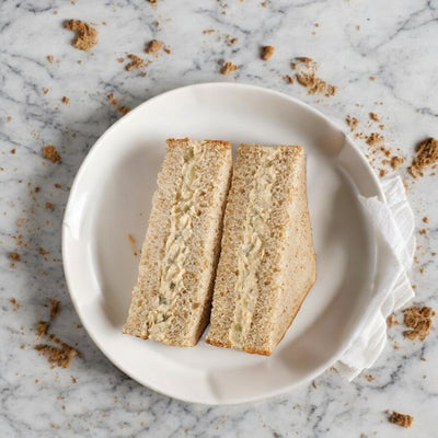 An E.A. Sween Albacore Tuna Salad Sandwich, halved and served on whole-grain bread with a creamy filling, placed on a white plate atop a marble surface with crumbs scattered around.