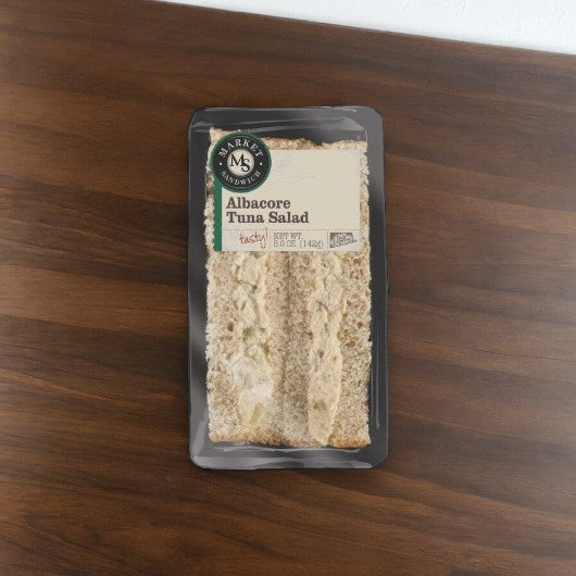 A packaged sandwich labeled "Albacore Tuna Salad Sandwich- 1 Count" on a wooden surface. The transparent packaging with a black border displays the sandwich and the brand name "E.A. Sween.