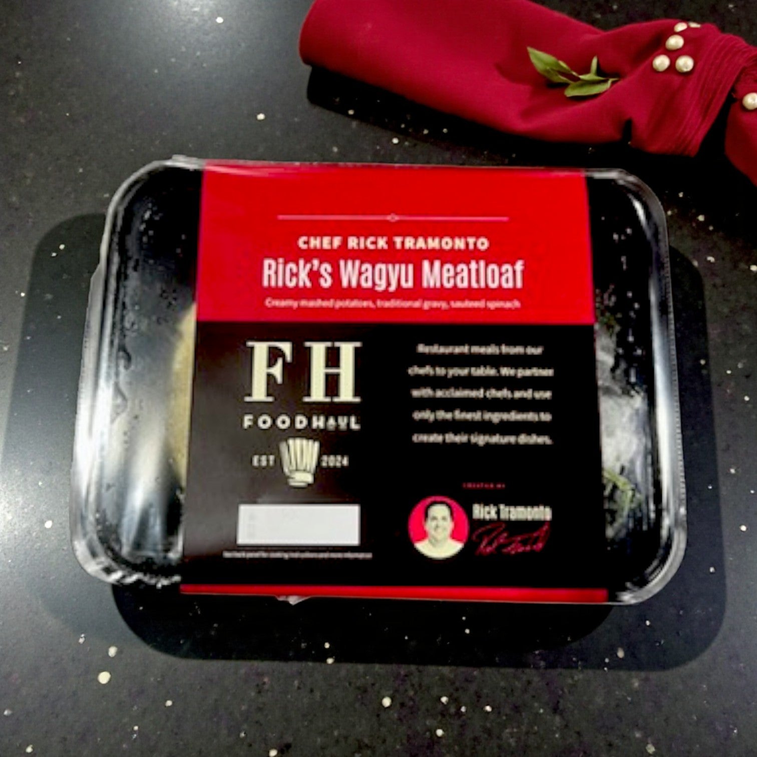 A FoodHaul Frozen Meal—Rick's Wagyu Meatloaf by Chef Rick Tramonto rests on a dark countertop beside a folded red napkin with gold and pearl accents.