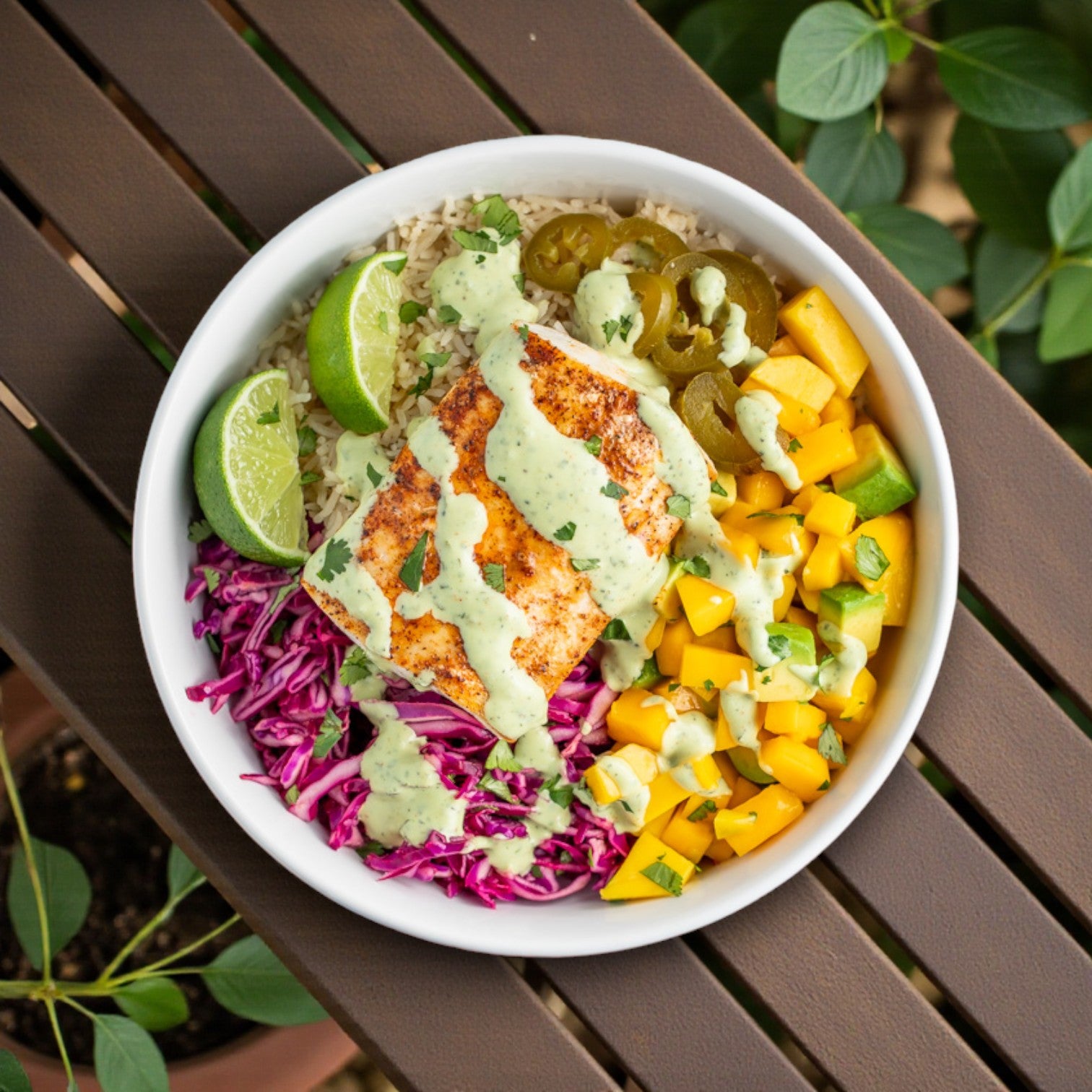 A white bowl with rice, grilled Alfa International Mahi Mahi Portions, red cabbage, mango, jalapeños, lime wedges, and green sauce sits on a brown slatted table with green foliage in the background. Ideal for featuring Alfa International seafood.