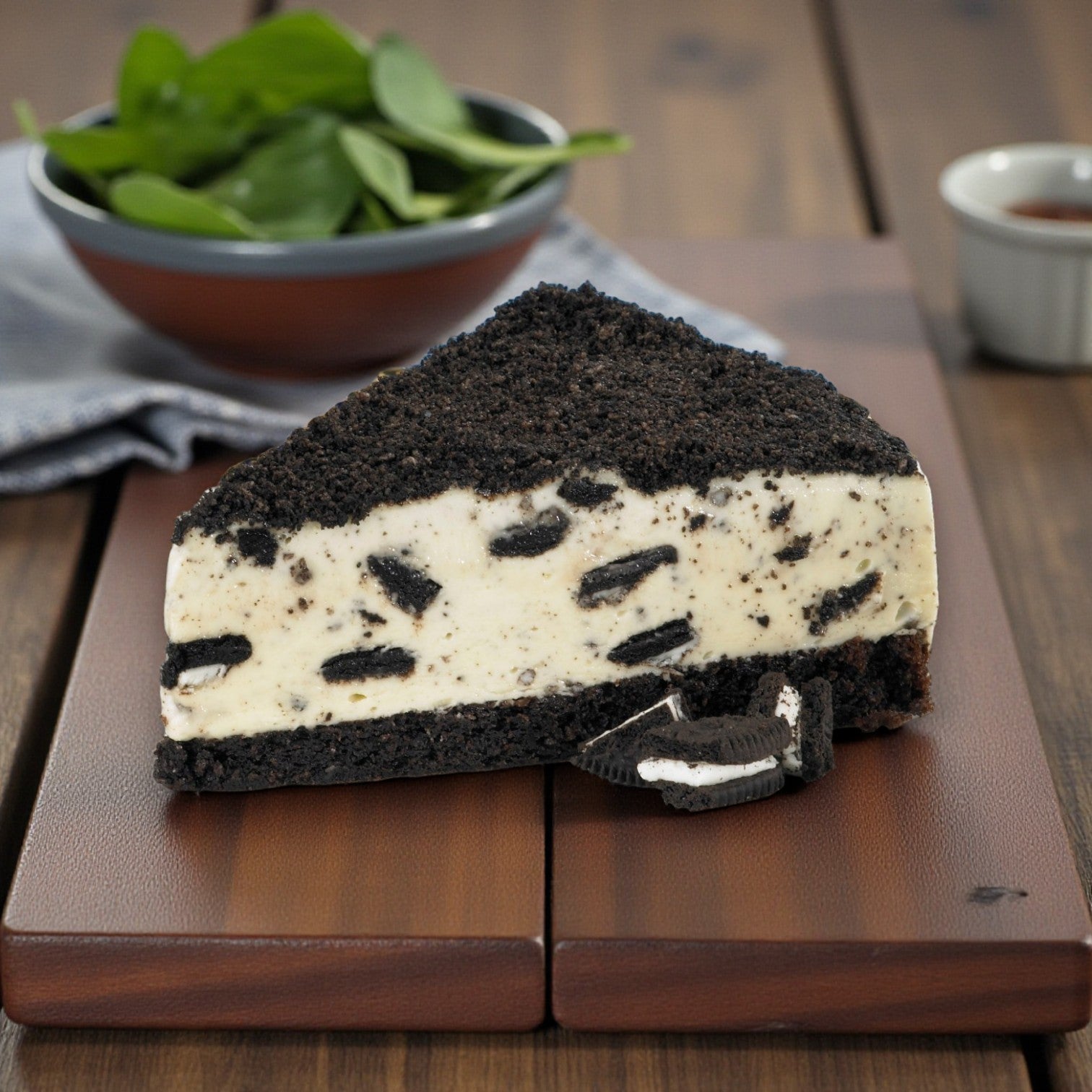 A slice of Easy Lunches’ Frozen Chef’s Quality – Oreo Mousse Cake – 14 Slices with an Oreo crust rests on a wooden board, making it a tempting pre-sliced dessert for cookies & cream mousse fans.