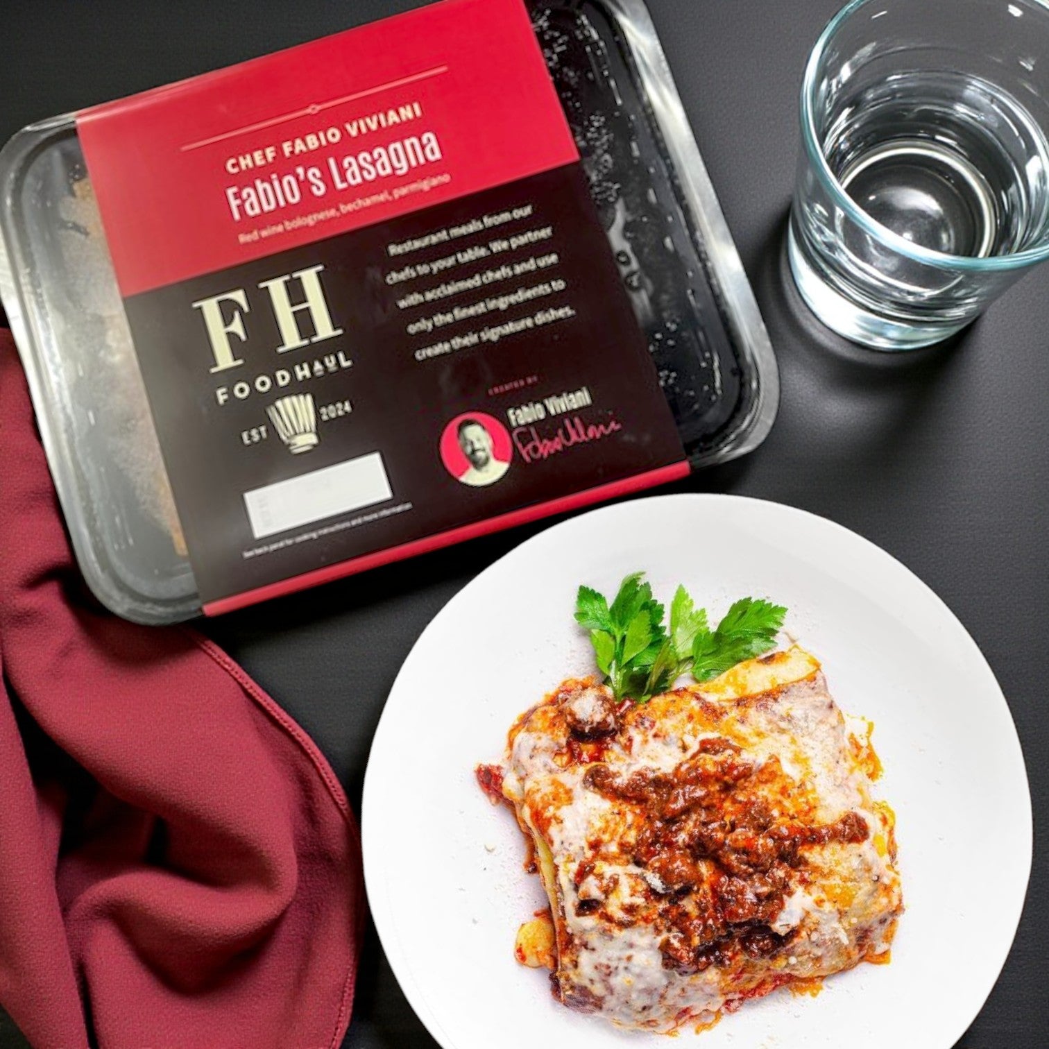 A serving of lasagna with parsley garnish, water, and a maroon napkin beside FoodHaul Frozen Meals- Signature Chef Meal Combo—5 Gourmet Entrées, bringing restaurant-quality flavor to your table.