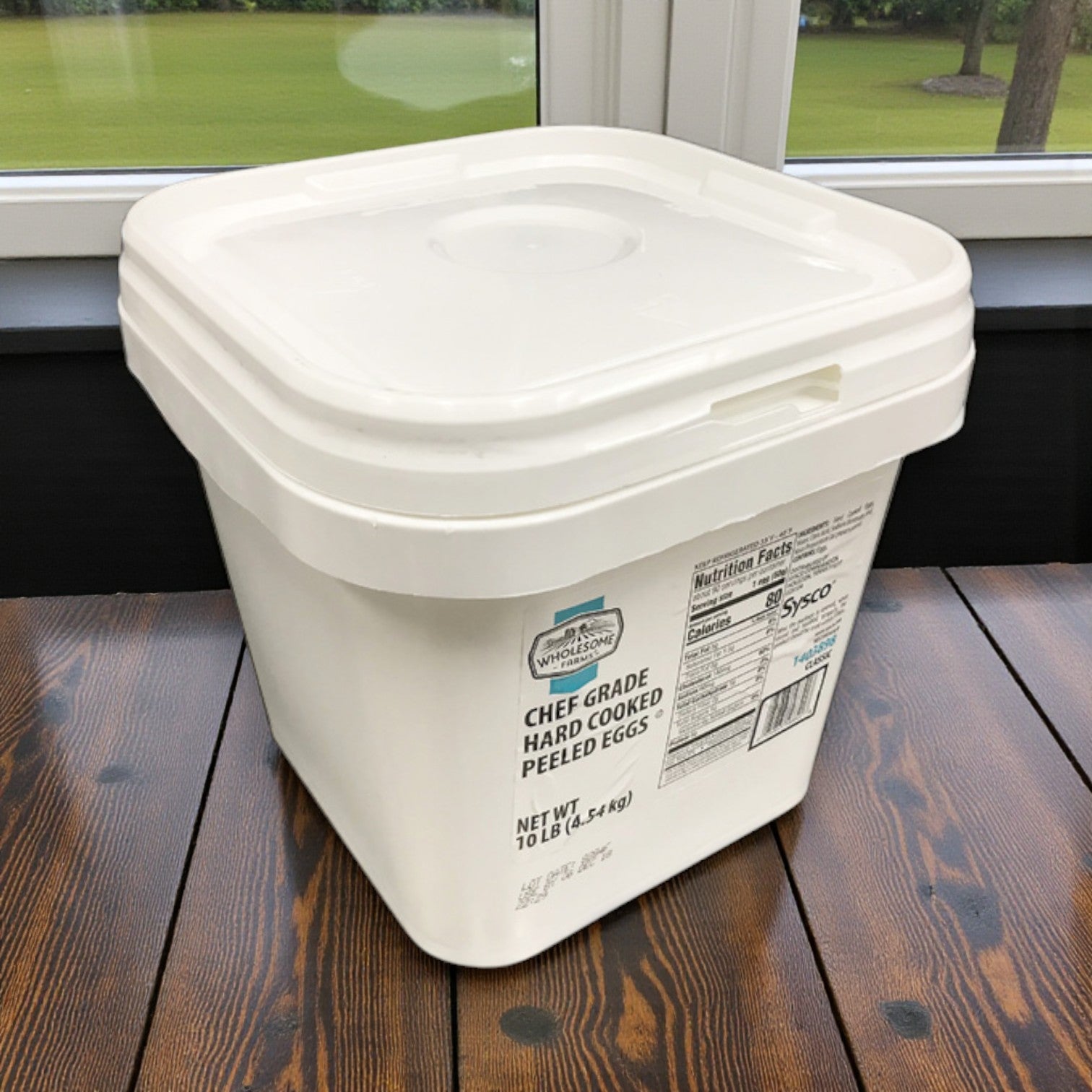 A large white Sysco Wholesome Farms Classic container of hard-boiled whole peeled eggs (1/10 lb) sits on a wooden table by windows, ideal for foodservice bulk storage or ready-to-serve use.