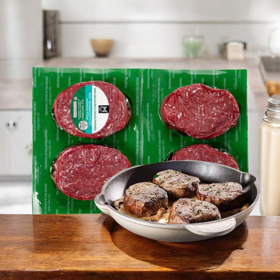 Four vacuum-sealed Member's Mark Grass Fed Tenderloin Beef Steaks (1 Pack, 4 steaks) are displayed behind a skillet with cooked pieces on a kitchen counter—ideal for elevating your gourmet dining experience.