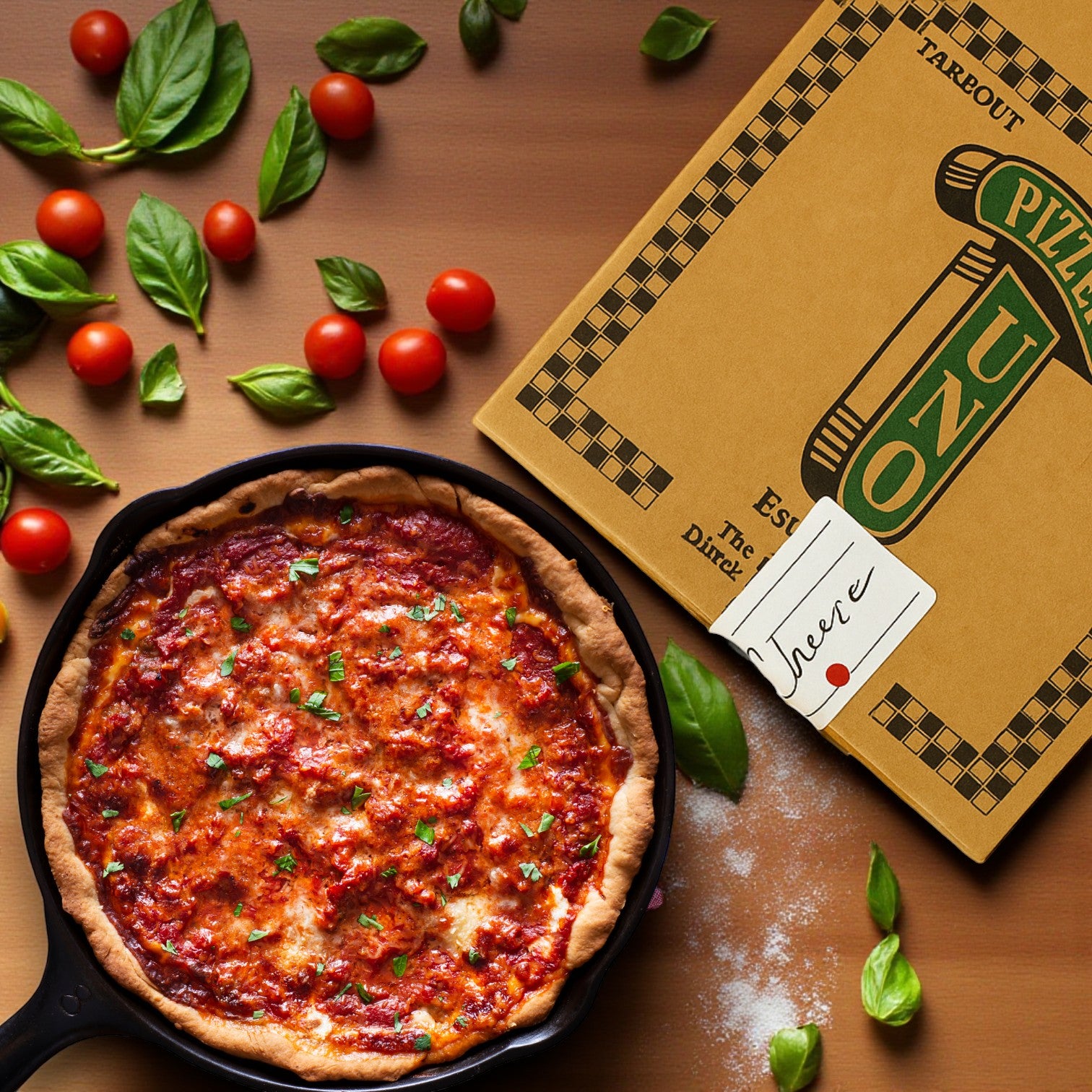 A 9” Uno’s Pizza Deep Dish Small Cheese Pizza (Frozen - Made in Restaurant) by Pizzeria Uno sits in a skillet beside its takeout box, surrounded by fresh basil and cherry tomatoes on a wooden surface.