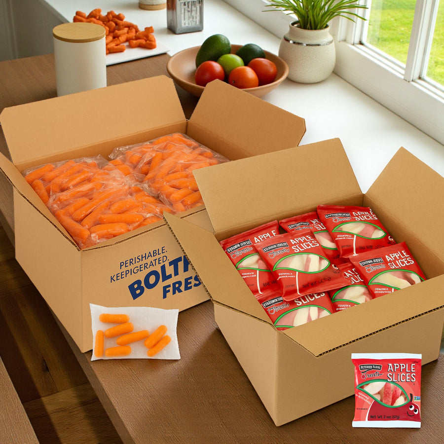 Two open Easy Lunches boxes on a kitchen counter: one with Fresh Sliced Apple Packs (2 oz., 100 packs), the other with Baby Carrots in Bags (1.6 oz., 200 bags); loose apples and carrots from the snack bundle are in the background.
