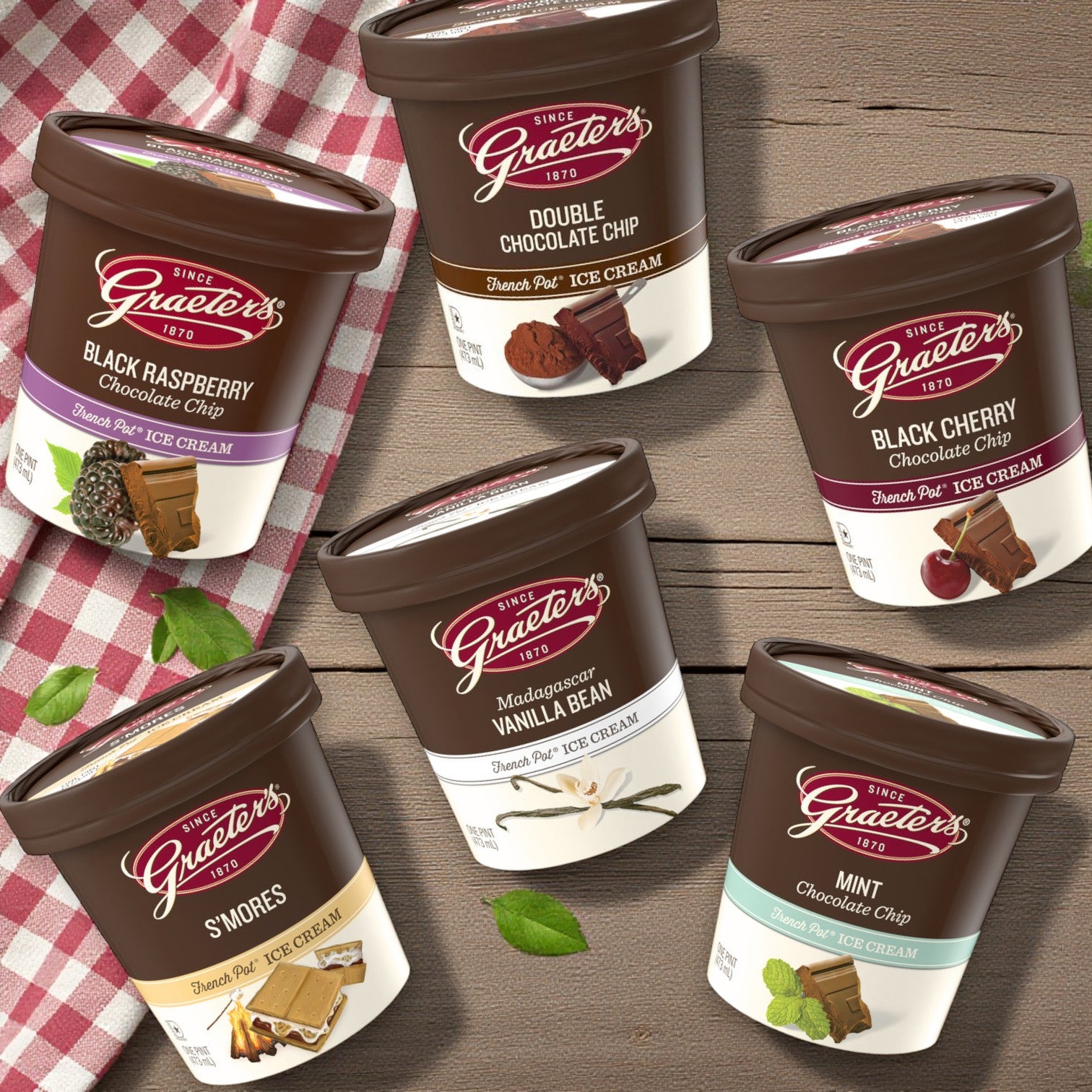 Six pints of Graeter’s Craft Ice Cream Combo Pack, featuring assorted flavors, are displayed on a wooden surface beside a red-and-white checkered cloth.