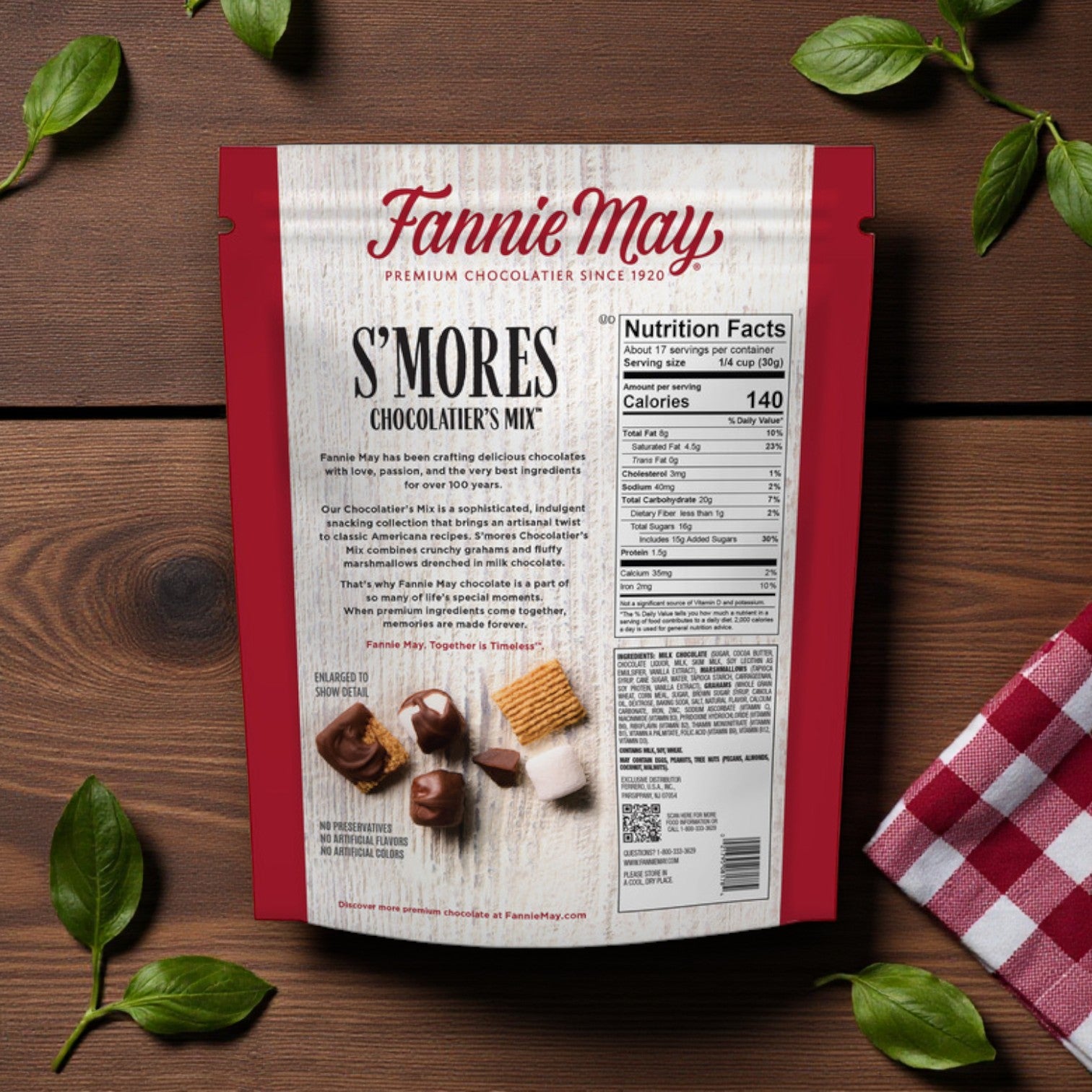 A Fannie May S’mores Snack Mix Bag, 18 oz, by Fannie May is shown on a wooden surface with green leaves and a red checkered cloth. The packaging displays nutrition facts and the product description.