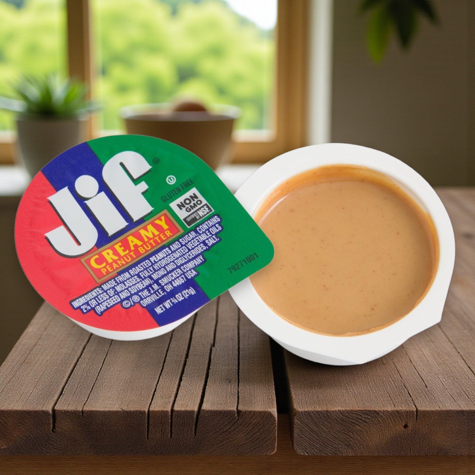 An open JIF Creamy Peanut Butter Cup (0.75 oz, 100 Count Case) rests on a wooden surface with blurred greenery visible through a window in the background.