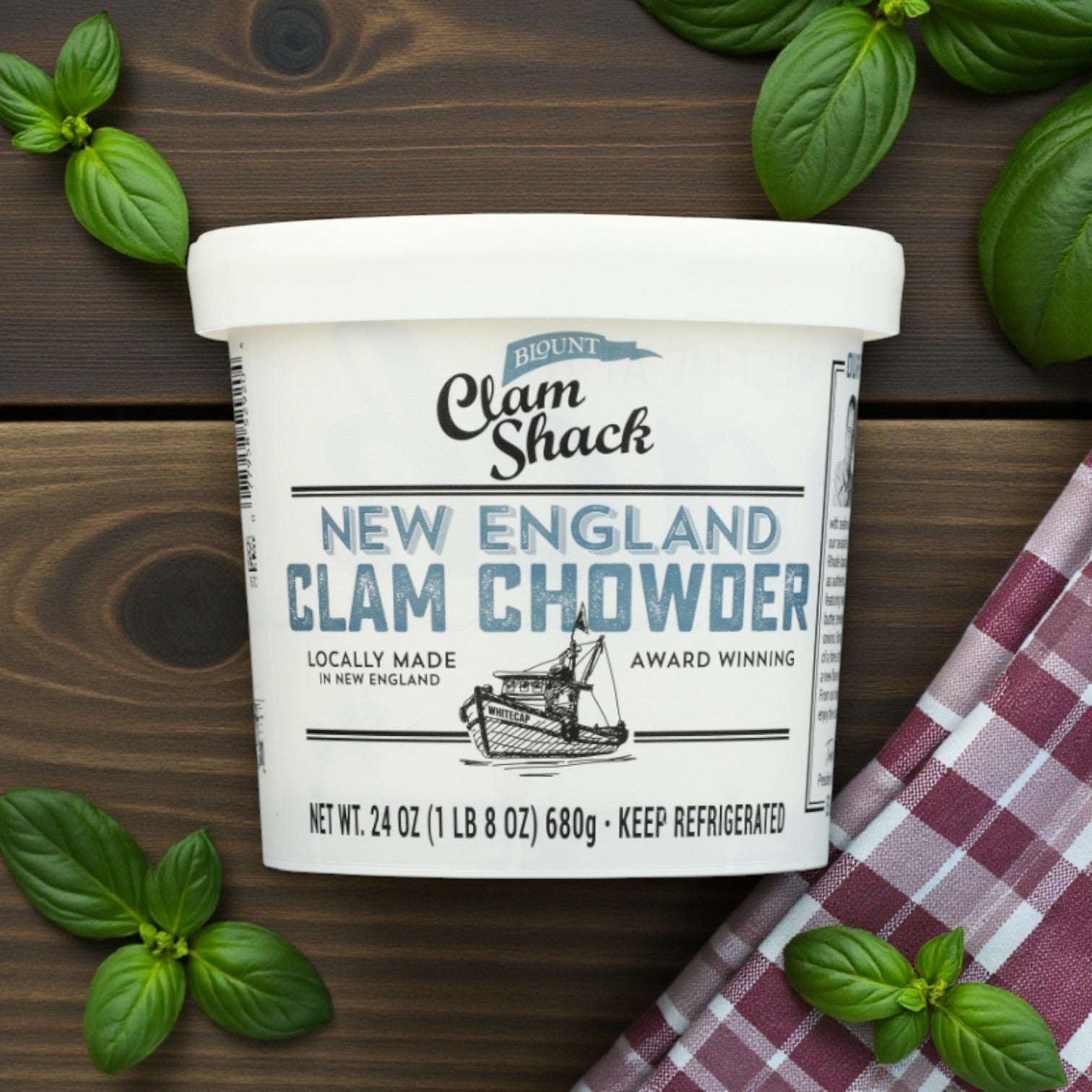 A 4-pack of Blount Fine Foods Clam Shack Premium Seafood Soup Combo (24 oz tubs) sits on a wooden surface with basil leaves and a red plaid cloth, the label highlighting its locally made, gourmet quality and award-winning flavors.