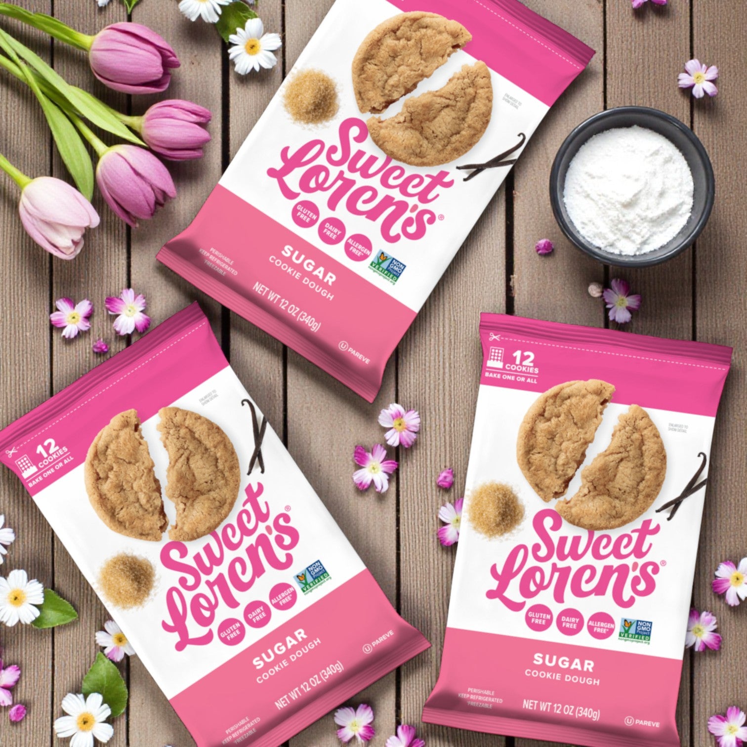 Three Sweet Loren’s Sugar Cookie Dough – Gluten-Free & Vegan 12 oz (3-Pack) packages are displayed on a wooden surface with flowers, a spoon, and a small bowl of flour.
