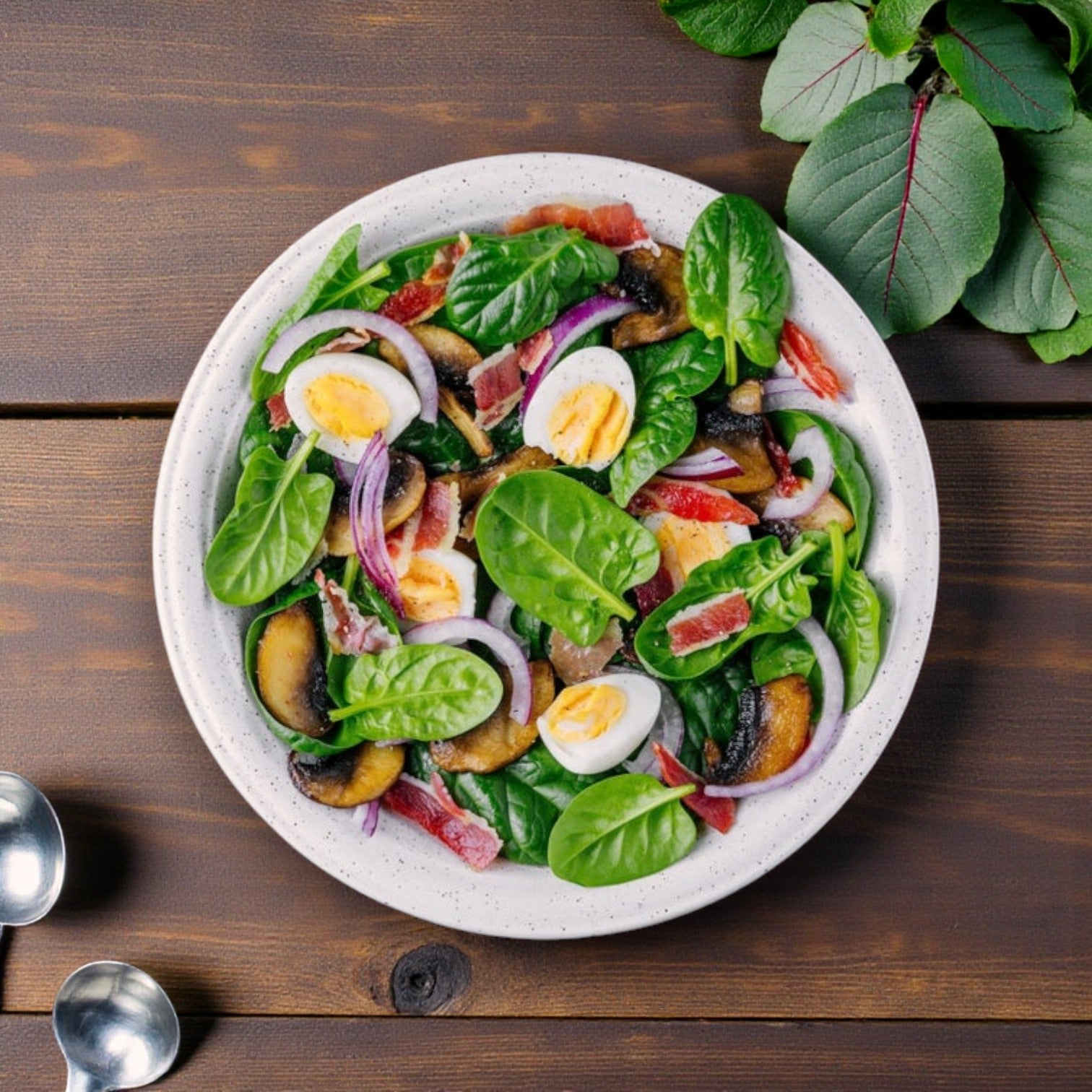 A white plate of spinach salad with Sysco Wholesome Farms Imperial Cage-Free Hard-Cooked Eggs, red onion, grilled mushrooms, bacon, and tomato sits on a wooden table among fresh leafy greens.